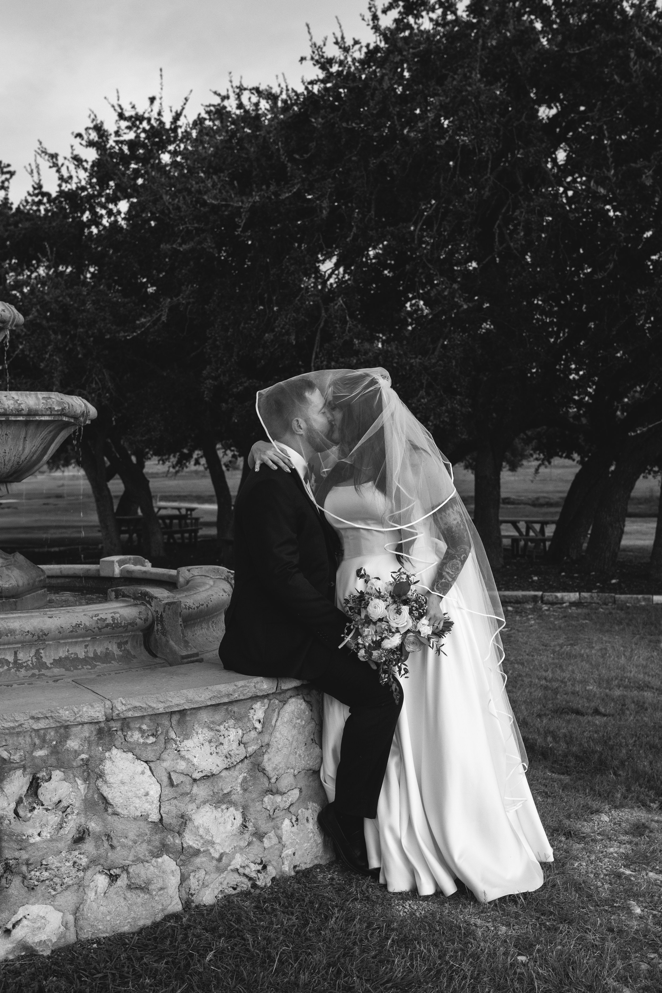 A bride and groom share a kiss outdoors during their wedding, with the bride wearing a white dress and a veil, the groom in a dark suit, holding a bouquet, and a fountain and trees in the background.