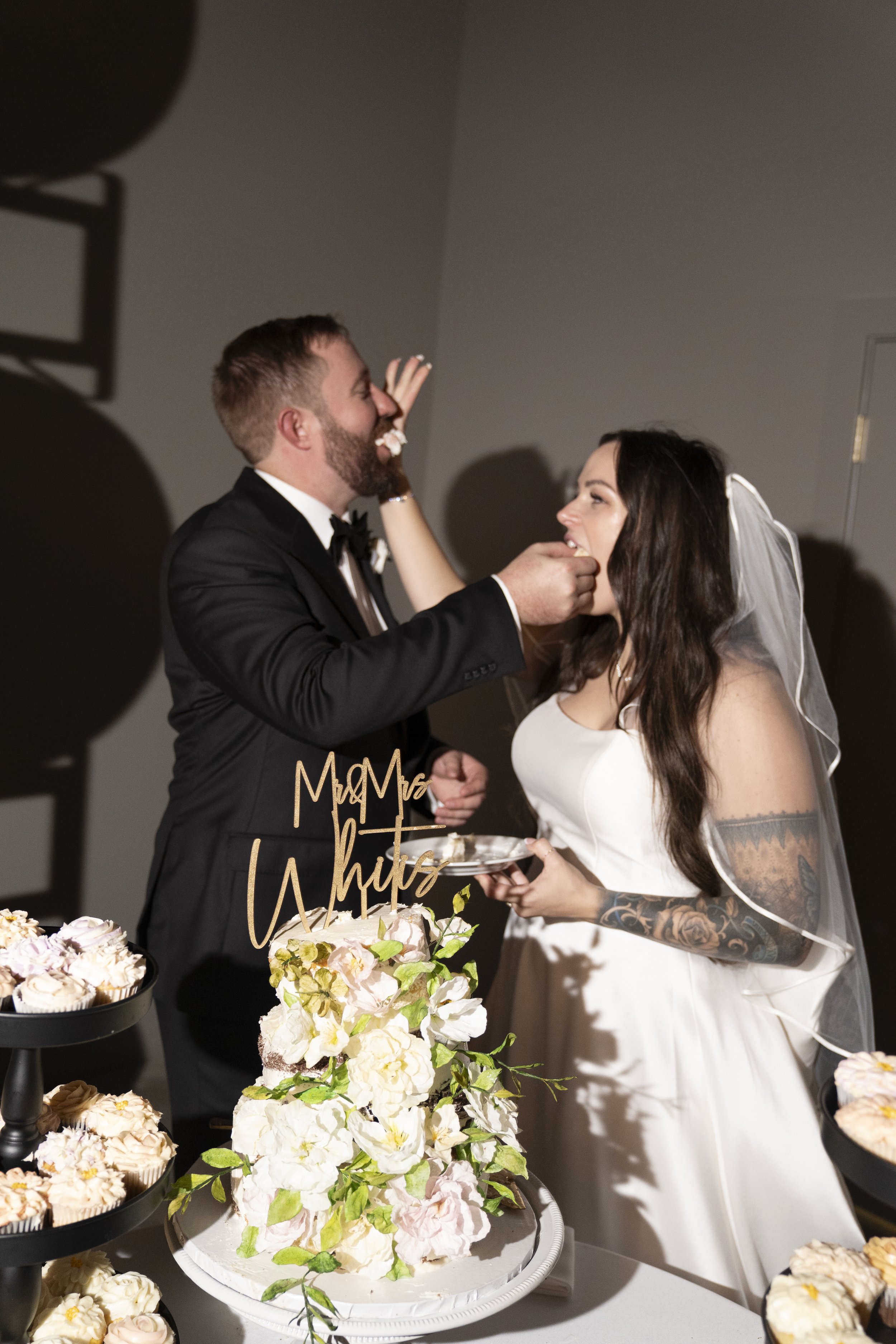 A newlywed couple feeds each other cake at their wedding reception with cupcakes and flowers on a table in front of them.