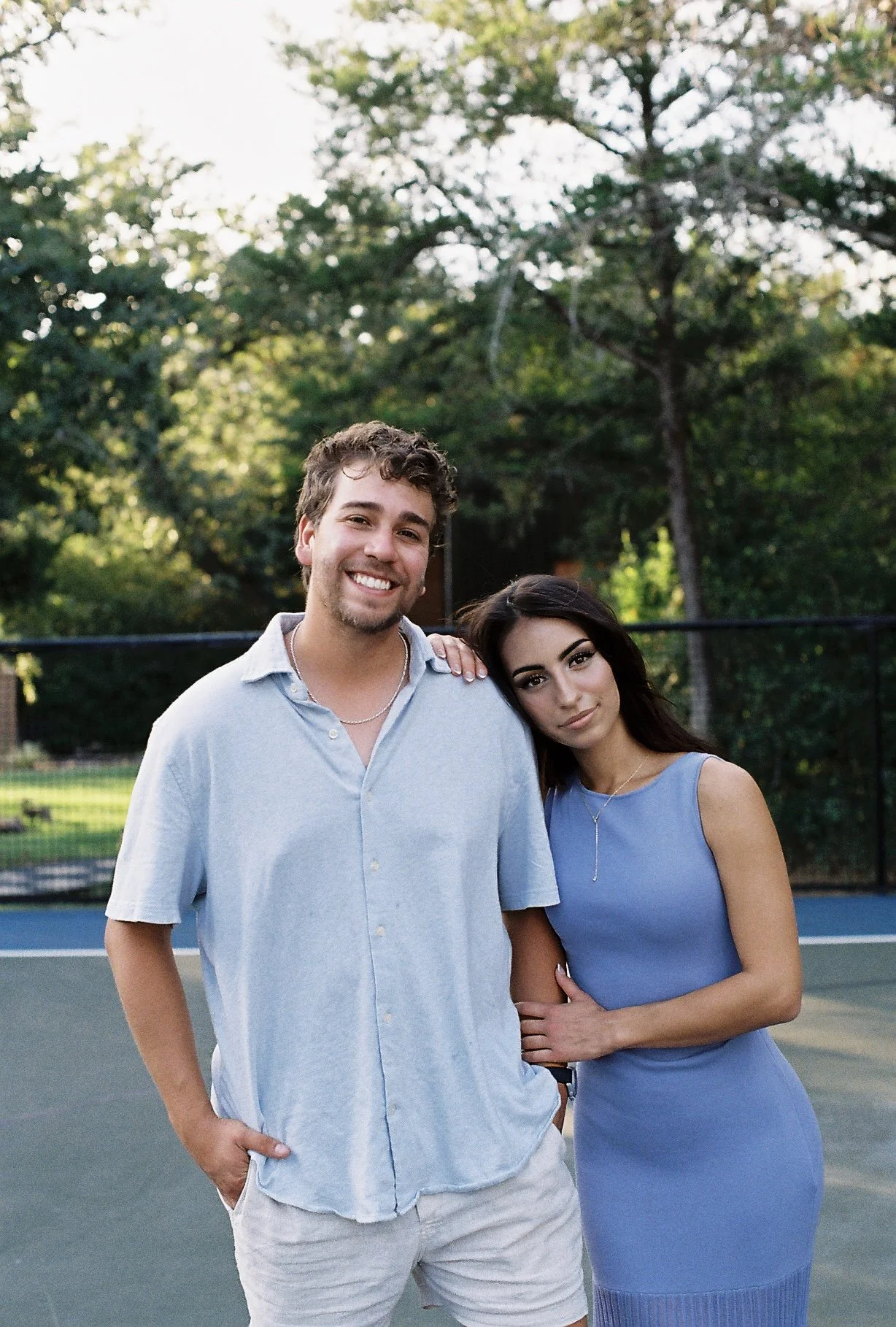 A smiling man and a woman standing on a tennis court. The man has dark wavy hair, light skin, a light blue shirt, white shorts, and a necklace. The woman has long dark hair, tan skin, a blue dress, and stands close to the man, leaning her head on his