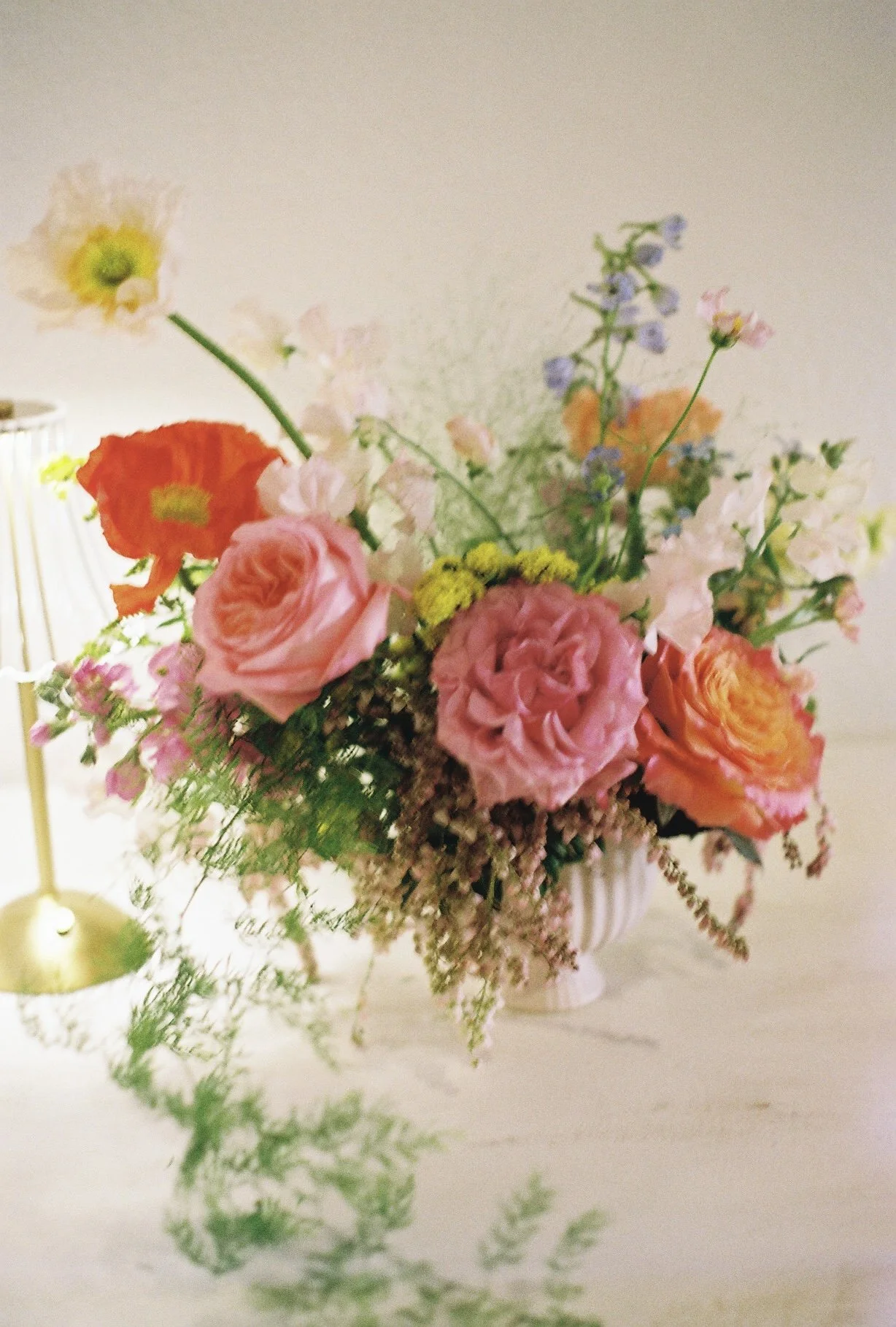 A colorful flower arrangement in a striped white vase on a white table. The bouquet includes pink roses, orange poppies, lavender, and other small flowers and greenery. A tall, gold lamp is partially visible on the left side of the image.