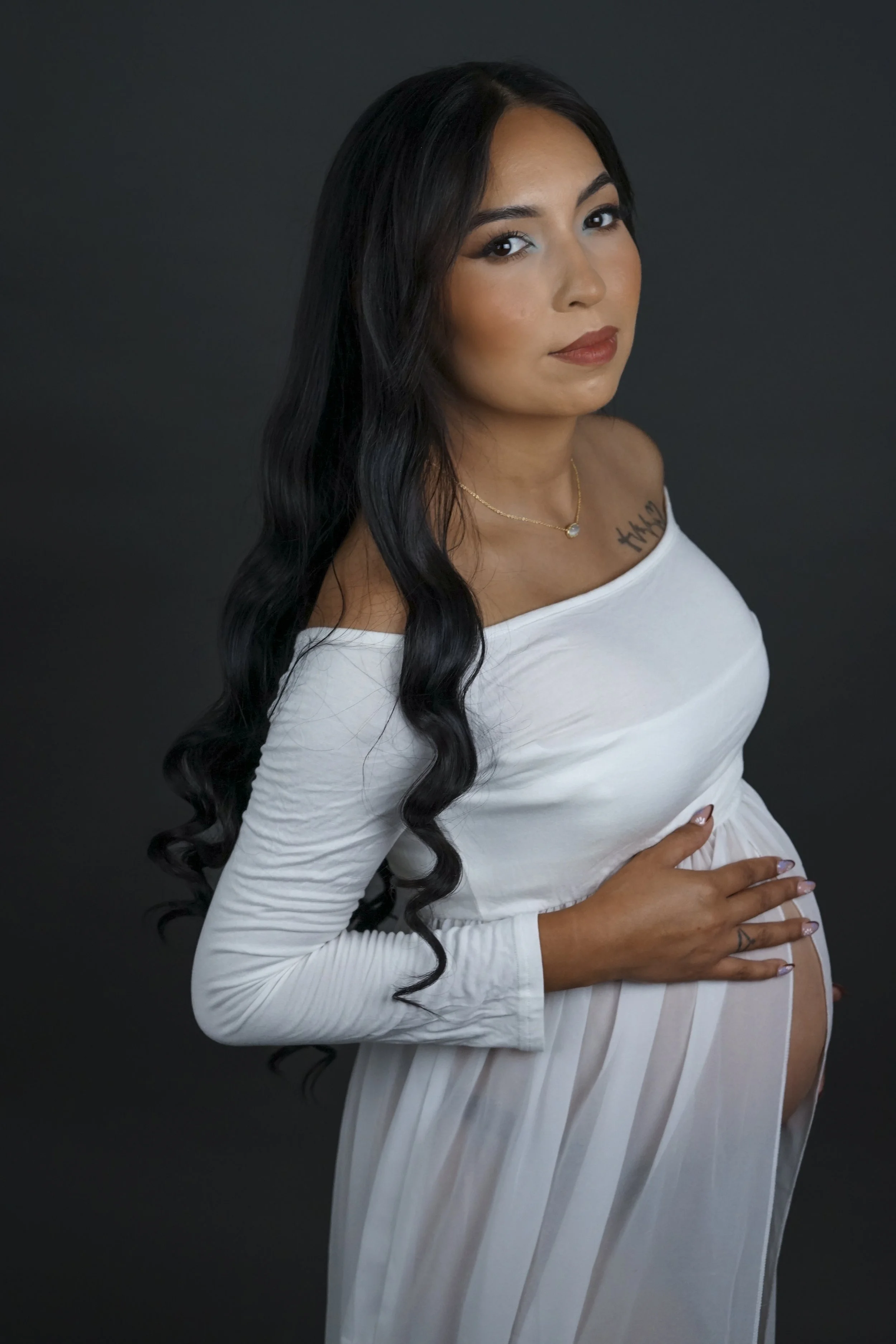 A pregnant woman with long black wavy hair wearing an off-the-shoulder white top and a matching flowing skirt, gently holding her belly against a dark background.