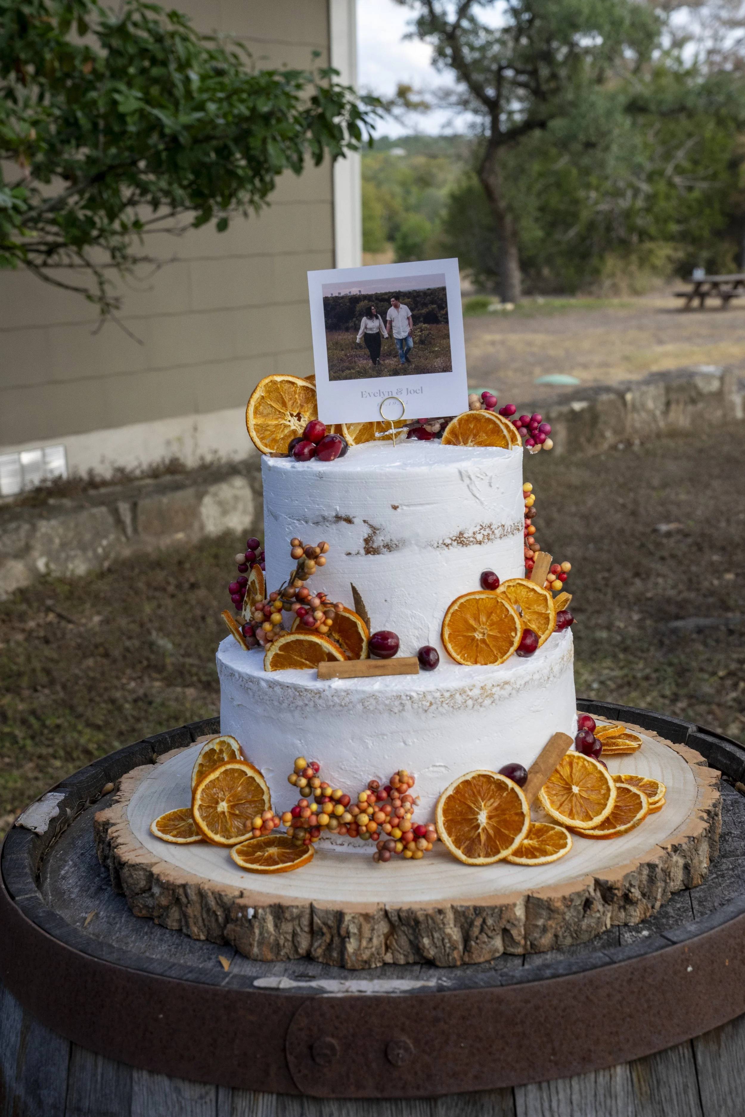 Two-tiered white wedding cake decorated with dried orange slices, small red berries, and cinnamon sticks, topped with a photo of a couple walking outdoors, on a wooden slab outside.
