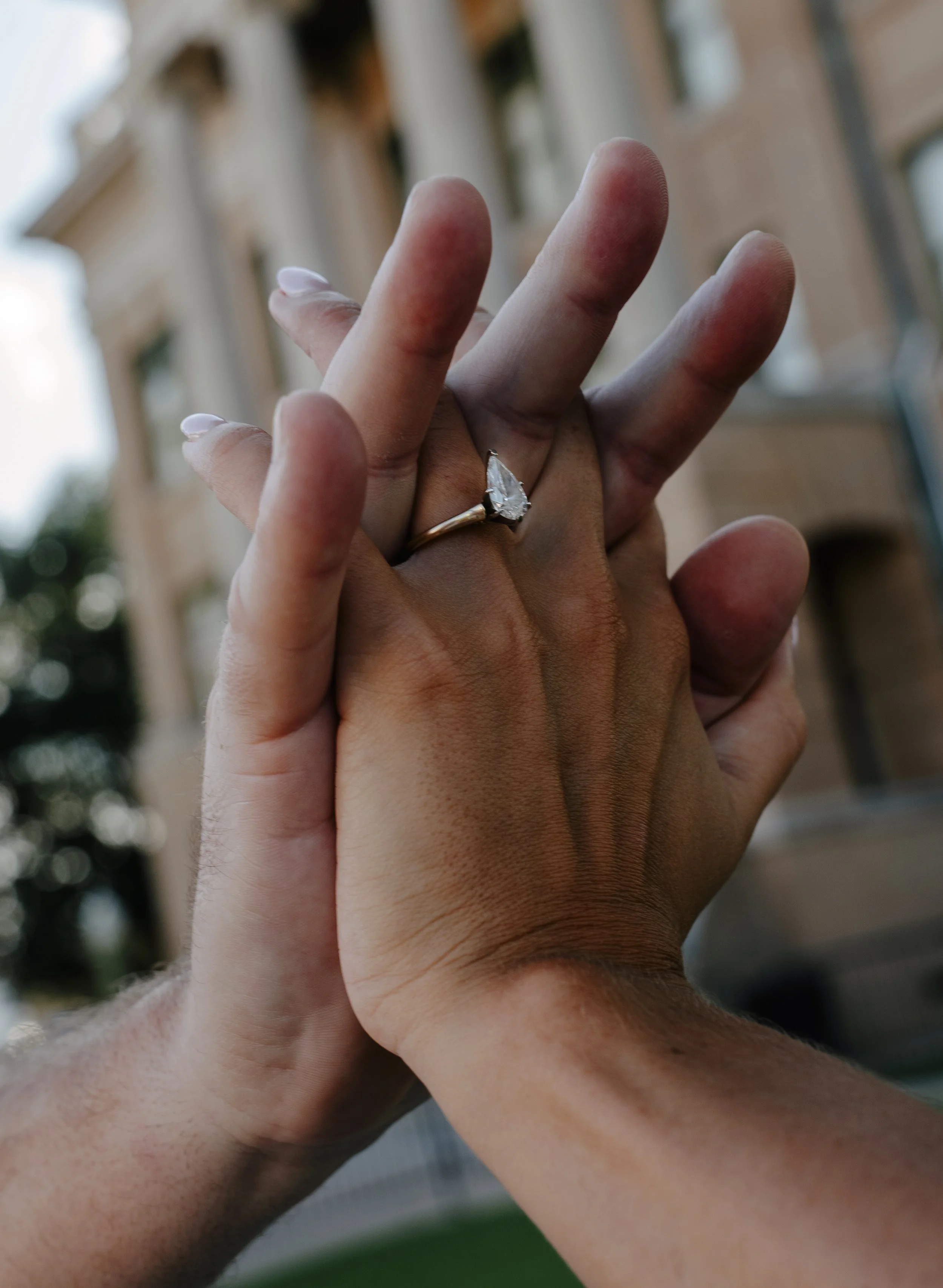 Close-up of two hands holding each other, one with a diamond engagement ring. The background shows blurred buildings and sky.