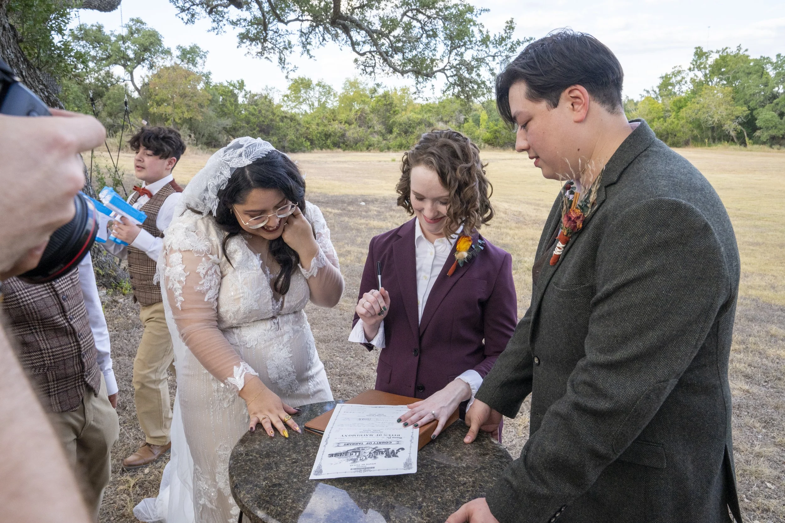 A group of people at an outdoor wedding ceremony, signing the marriage license, with a bride in a white lace dress and a groom in a grey suit.