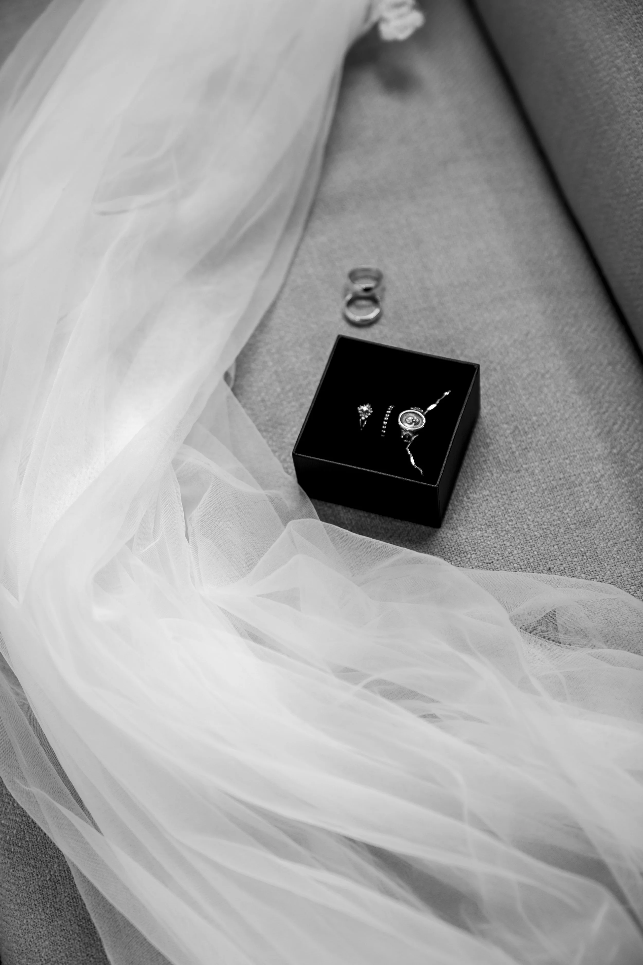 A black jewelry box containing a necklace, a ring, and earrings, placed on a fabric surface near a wedding veil, with a pair of rings nearby.