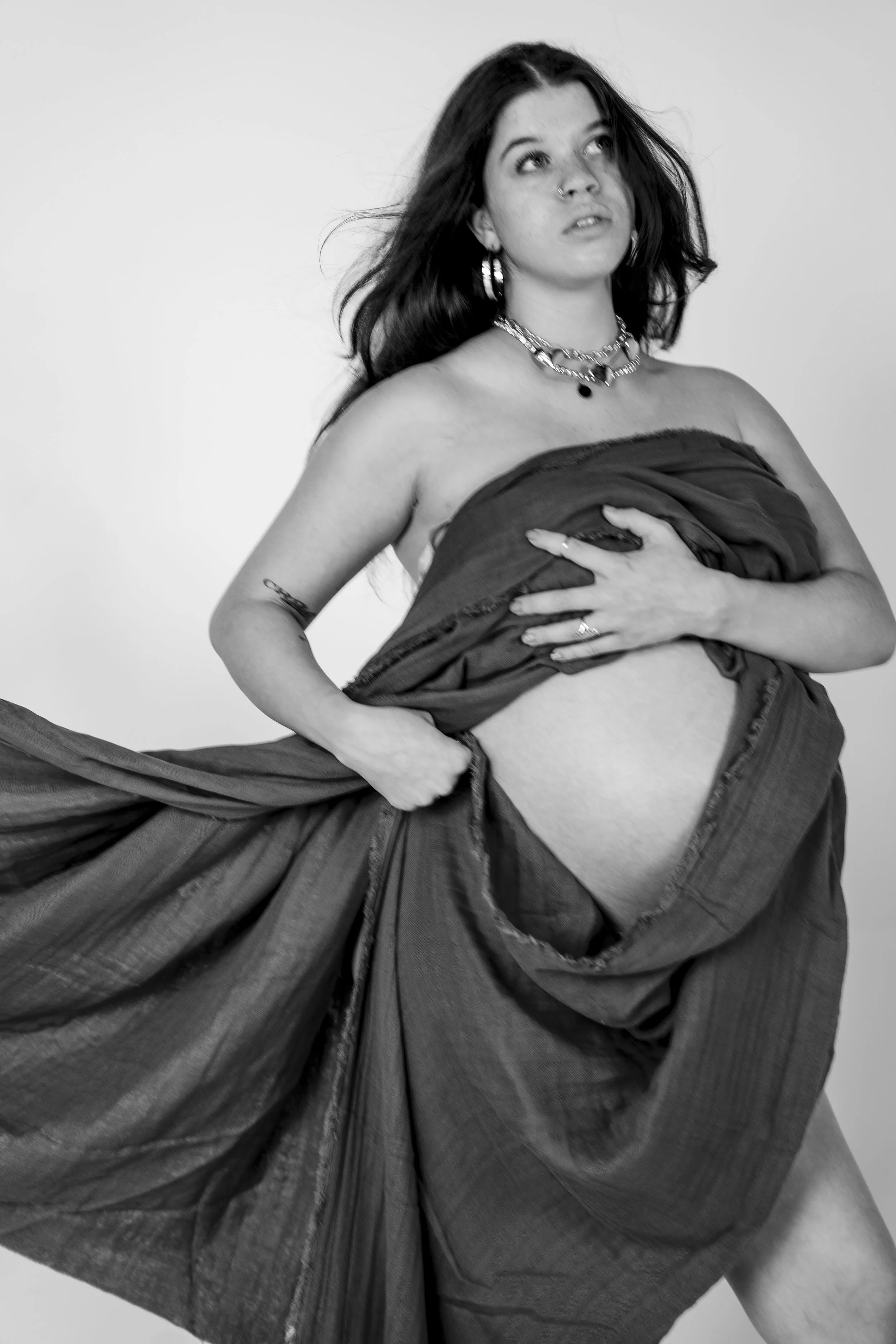 Black and white photo of a pregnant woman holding up her skirt, revealing her belly, in a studio setting.