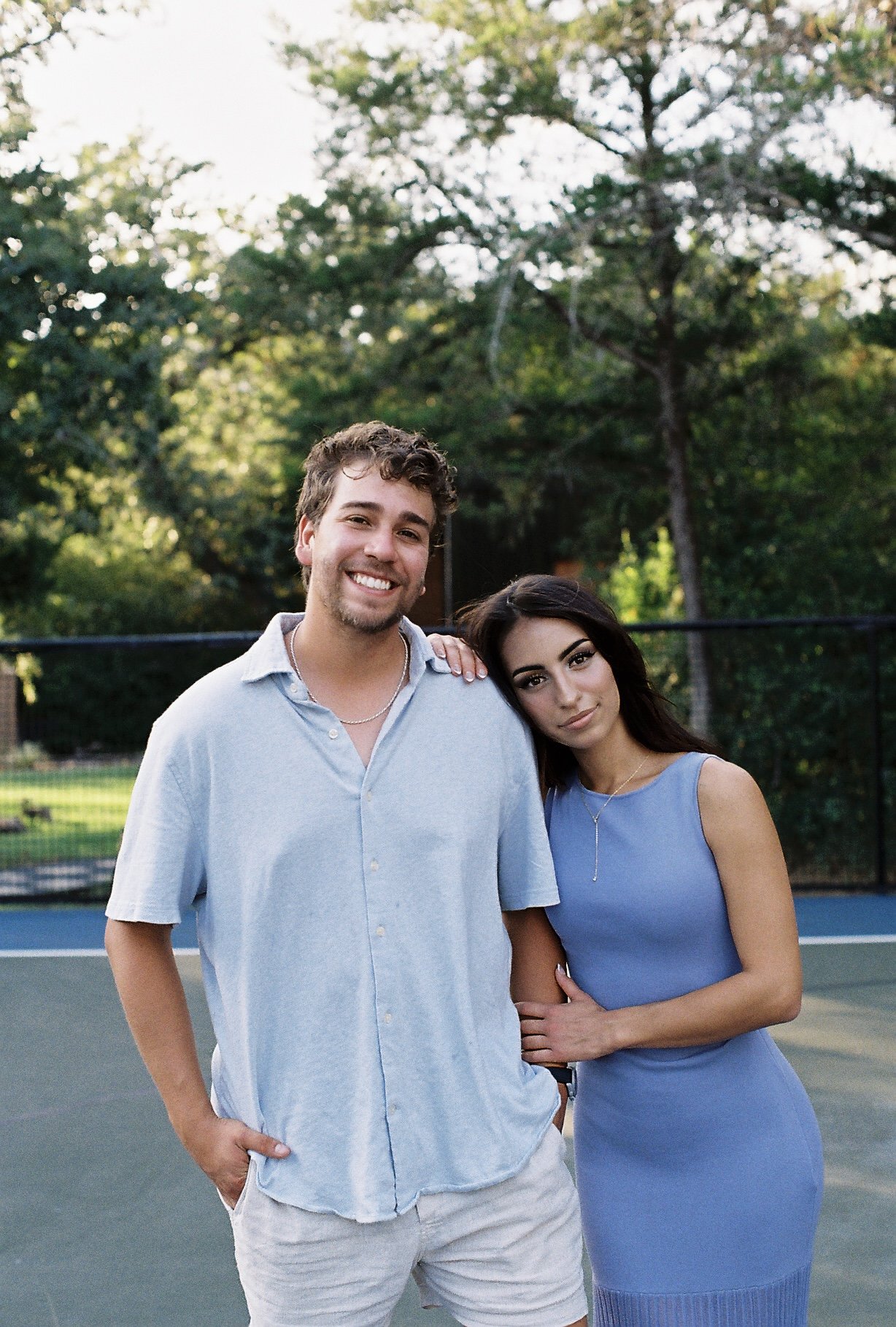 A young man and woman standing close together outdoors on a tennis court, smiling, with trees in the background.