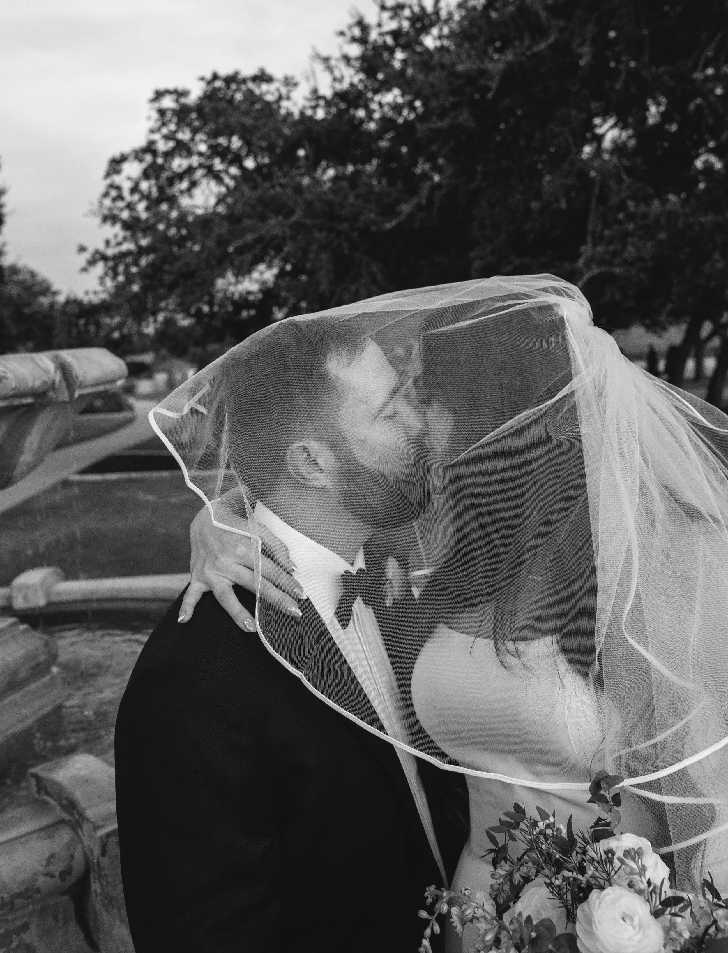 A bride and groom kissing outdoors, with the bride wearing a veil and holding a bouquet of flowers, during a wedding ceremony.
