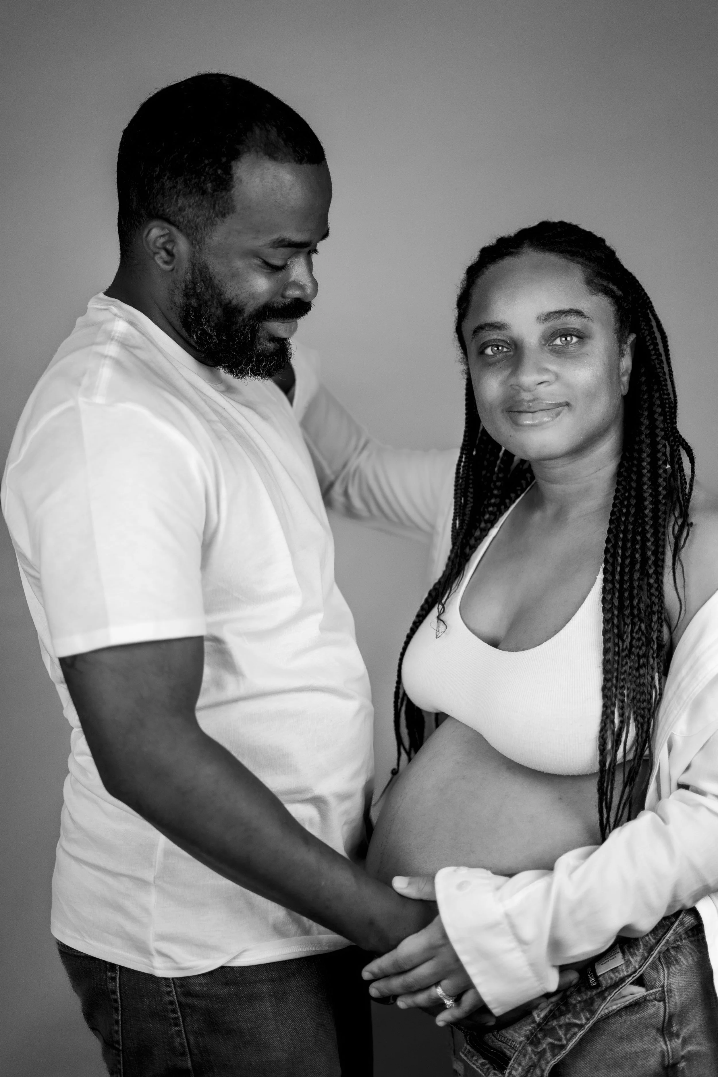 A black and white photo of a man and pregnant woman looking at each other. The man is wearing a white shirt, and the woman is wearing a white tank top, with her jacket off and an exposed pregnant belly.