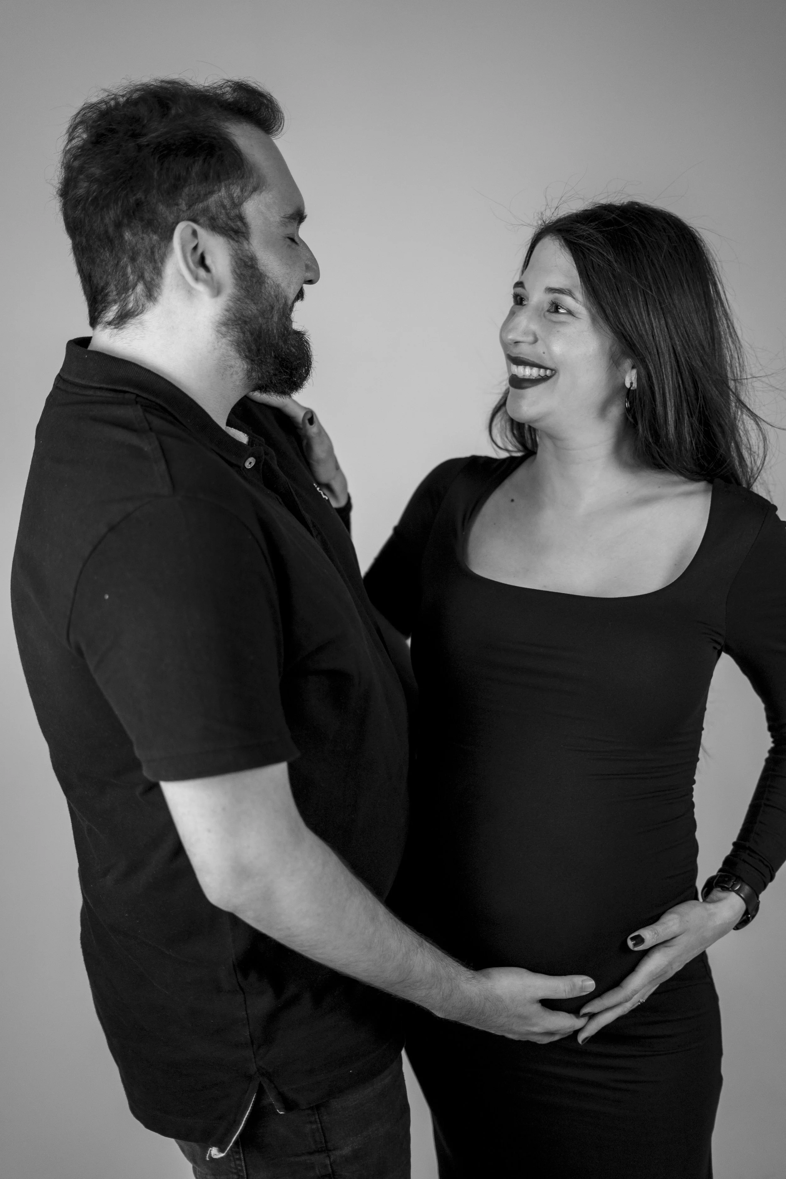 A man and pregnant woman smiling at each other, with the man's hand on her pregnant belly in a black and white photo.