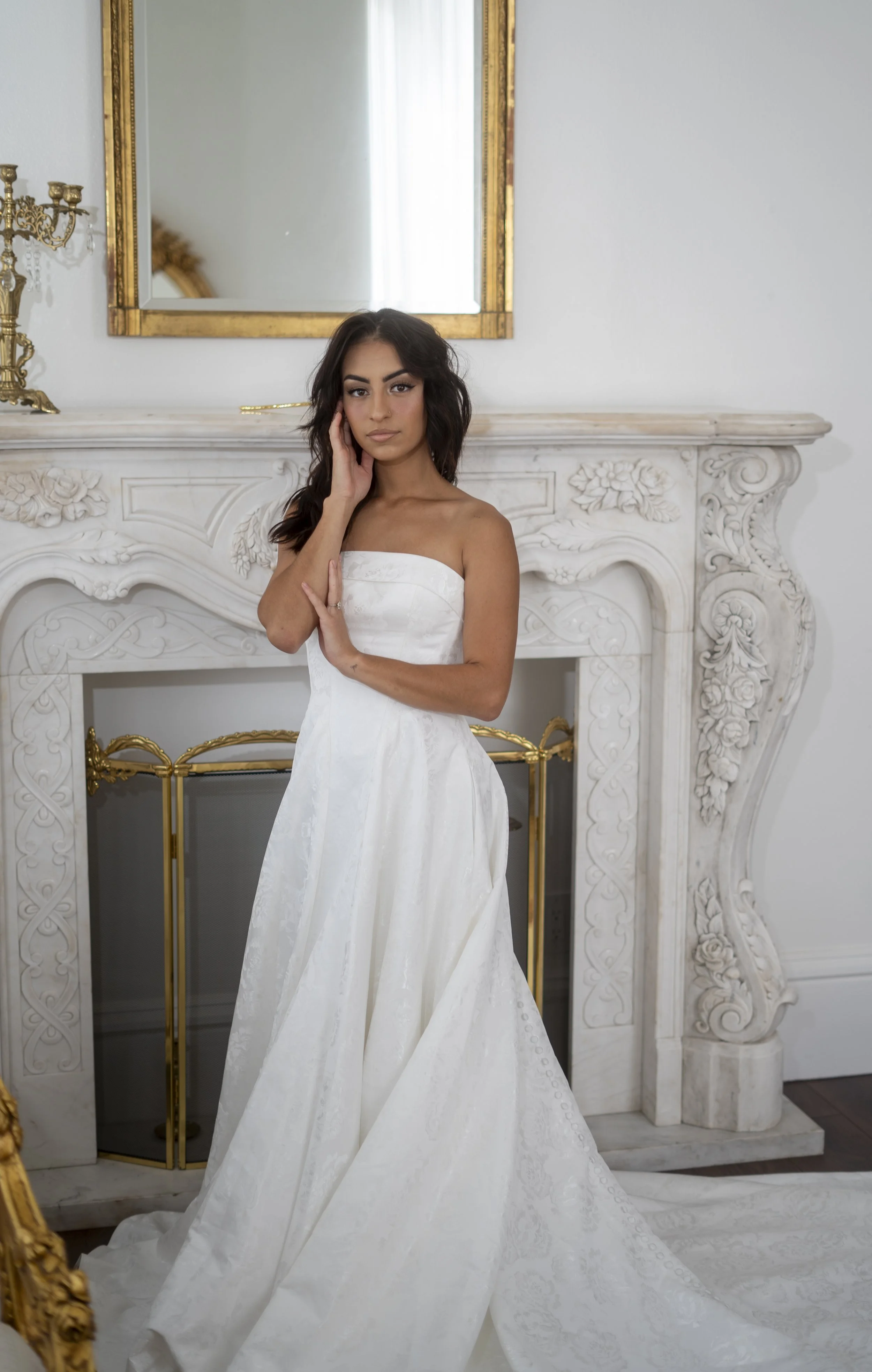 Woman in a white strapless wedding dress standing in front of a decorative white fireplace with floral carvings, a large mirror, and gold accents, touching her face with one hand and resting the other on her waist.