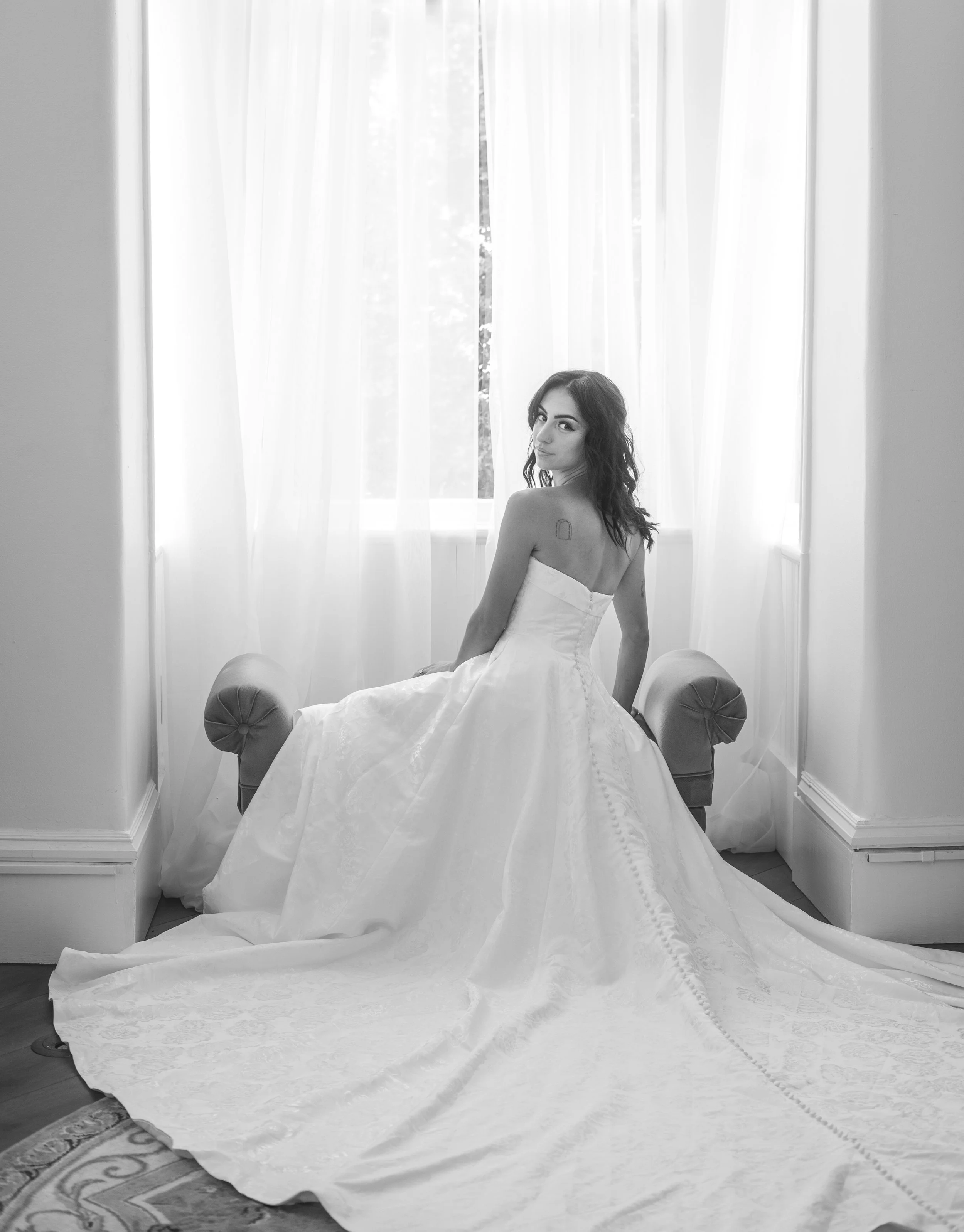 A woman in a wedding dress sitting on a window seat in front of tall, white, semi-transparent curtains, with a view of greenery outside.