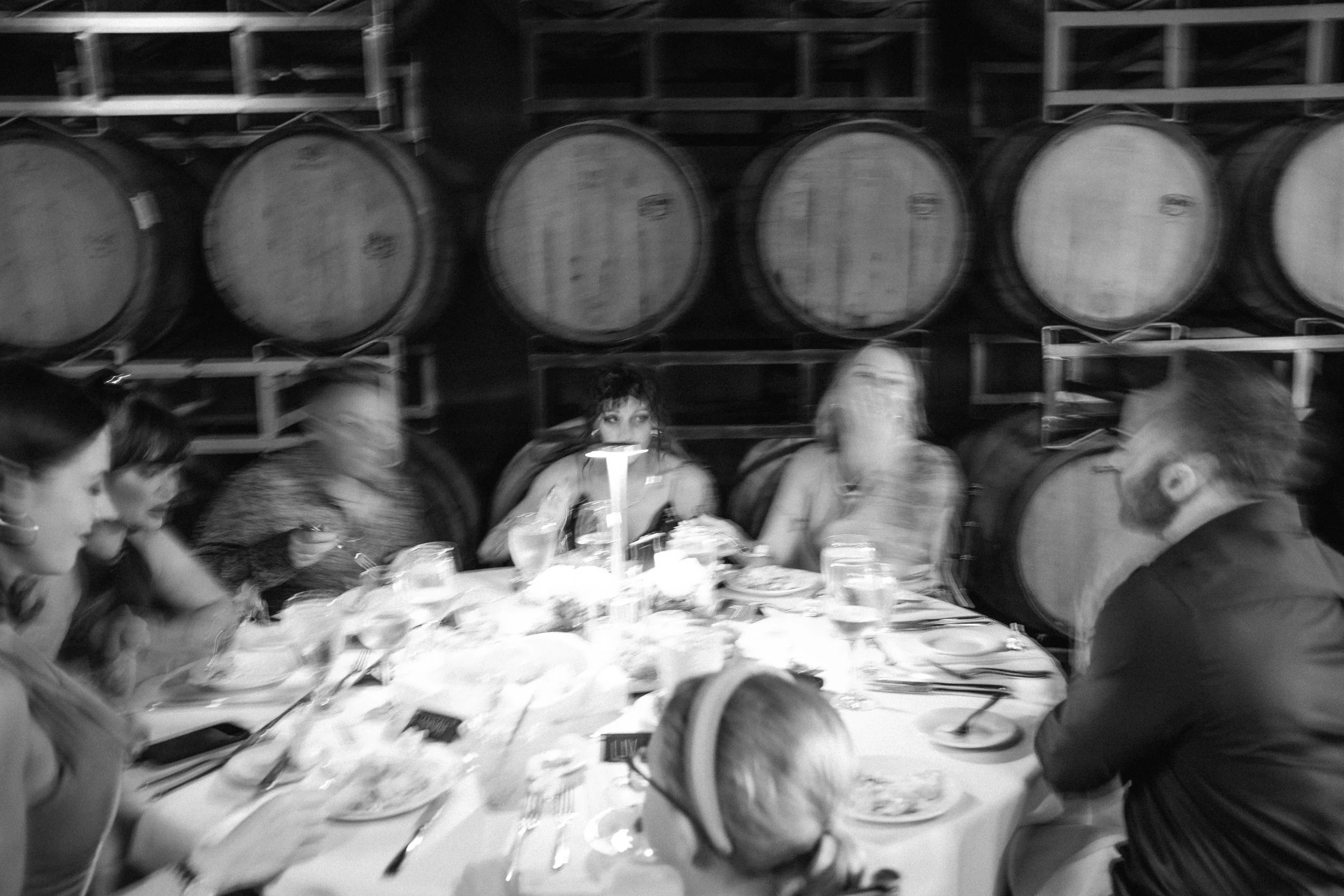 People gathered around a dining table with food, drinks, and tableware, in a setting with large barrels stored on racks in the background, in black and white.