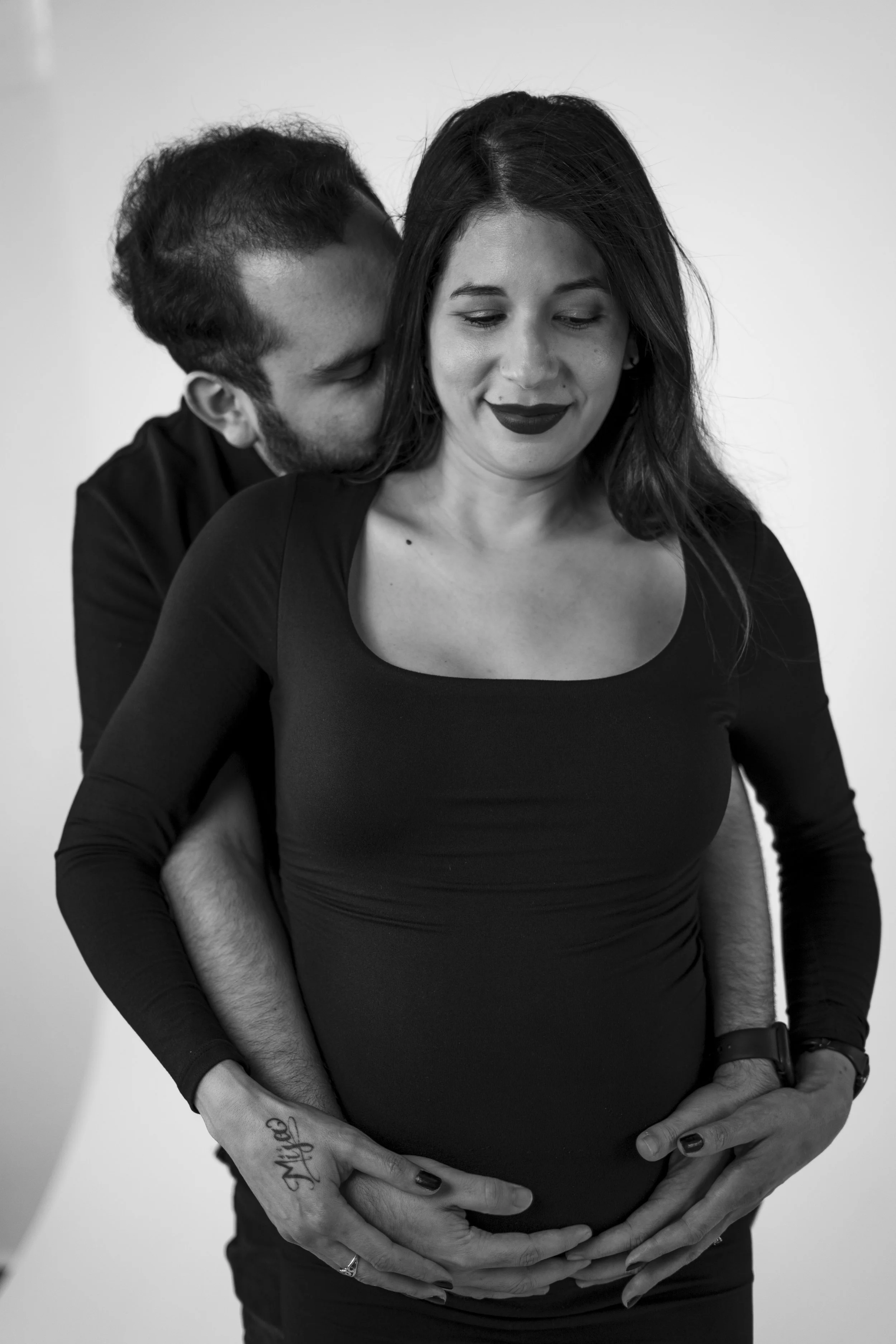 A man and a woman, with the man kissing the woman's neck from behind, and the woman's hands resting on her pregnant belly.