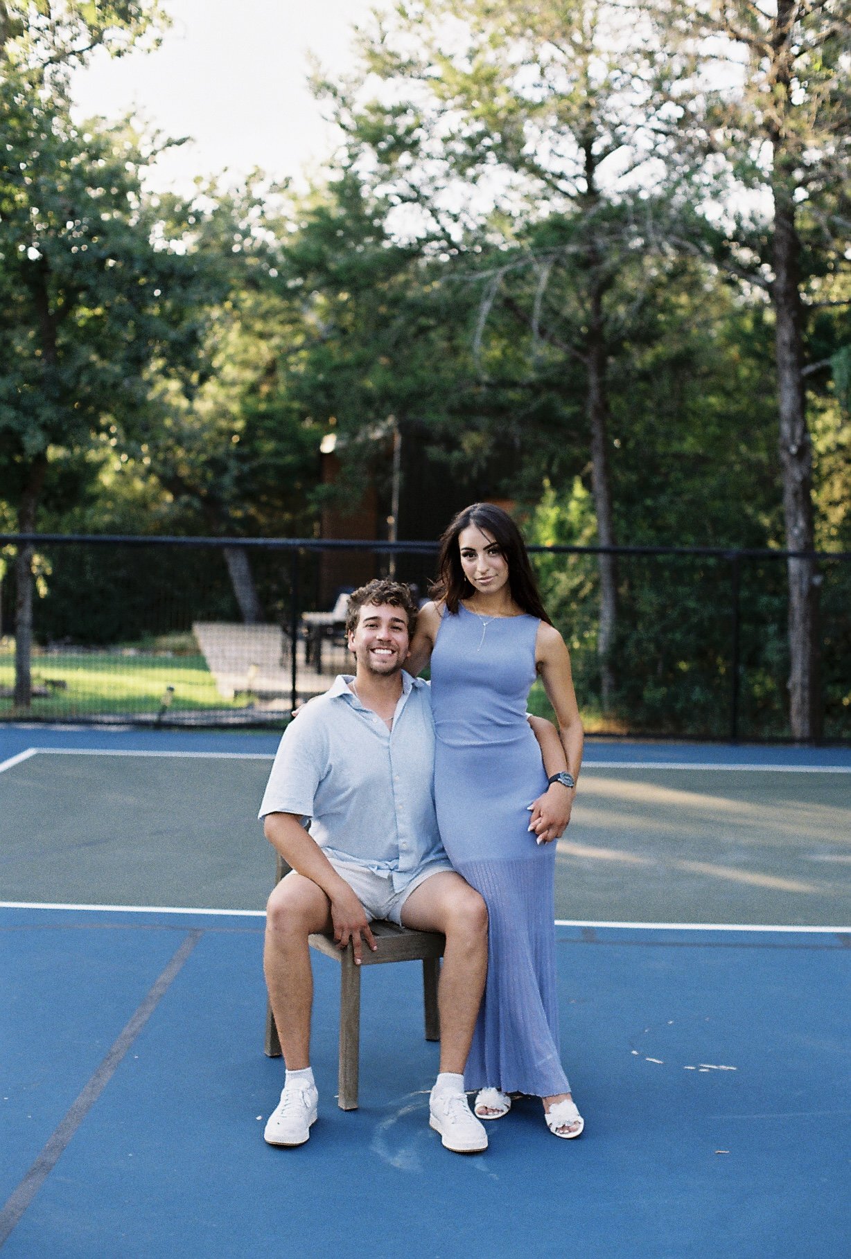 A man and woman posing on a tennis court. The man is sitting on a stool, smiling, wearing a light blue shirt, beige shorts, and white shoes. The woman is standing beside him, wearing a long, sleeveless blue dress and white sandals. They are surrounde