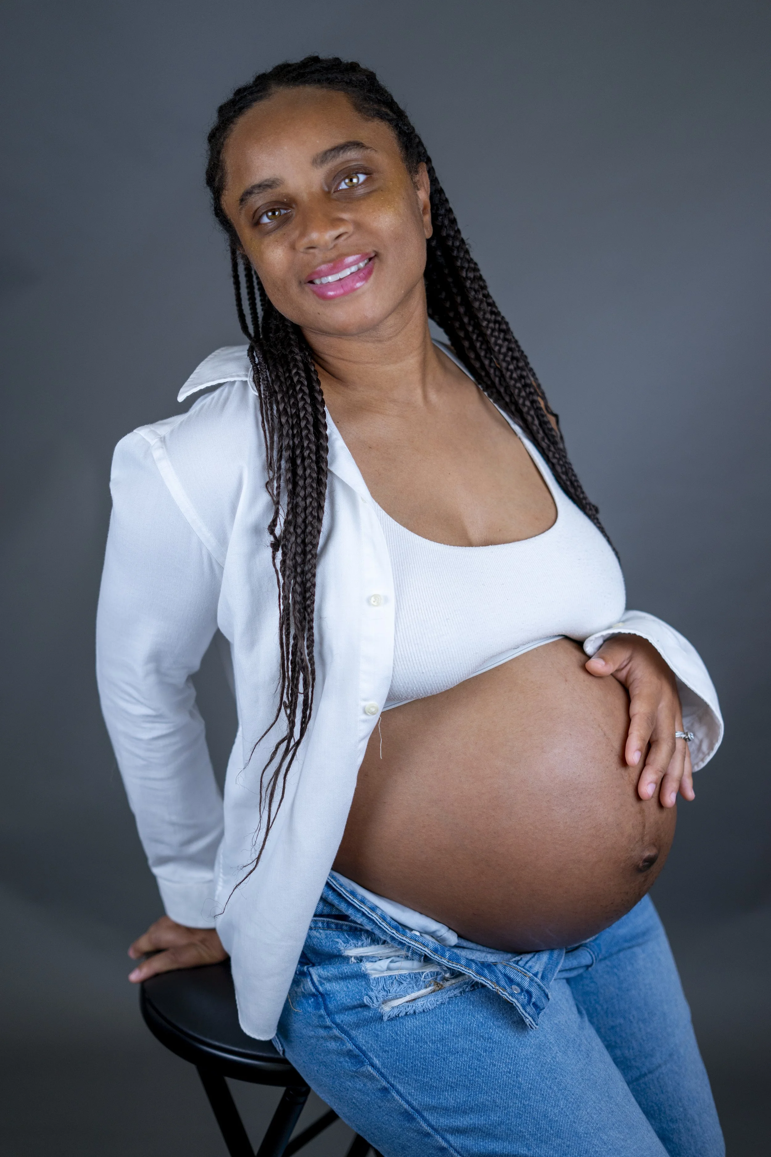 A pregnant woman with long braided hair smiling while sitting on a stool against a dark gray background, wearing a white tank top, unbuttoned white shirt, and ripped blue jeans.