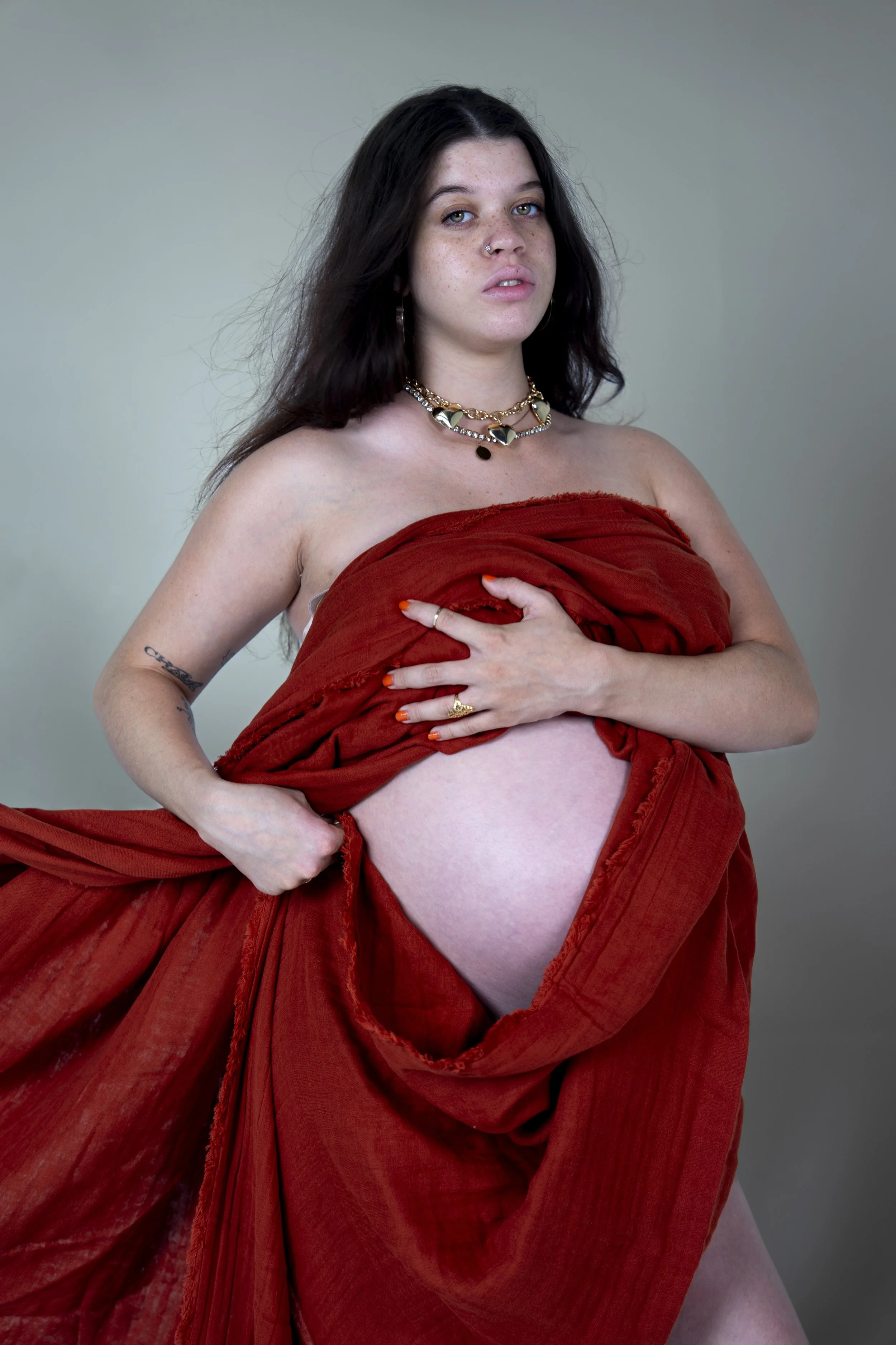 A woman with dark, wavy hair and freckles poses topless, draped in red fabric, against a plain gray background.