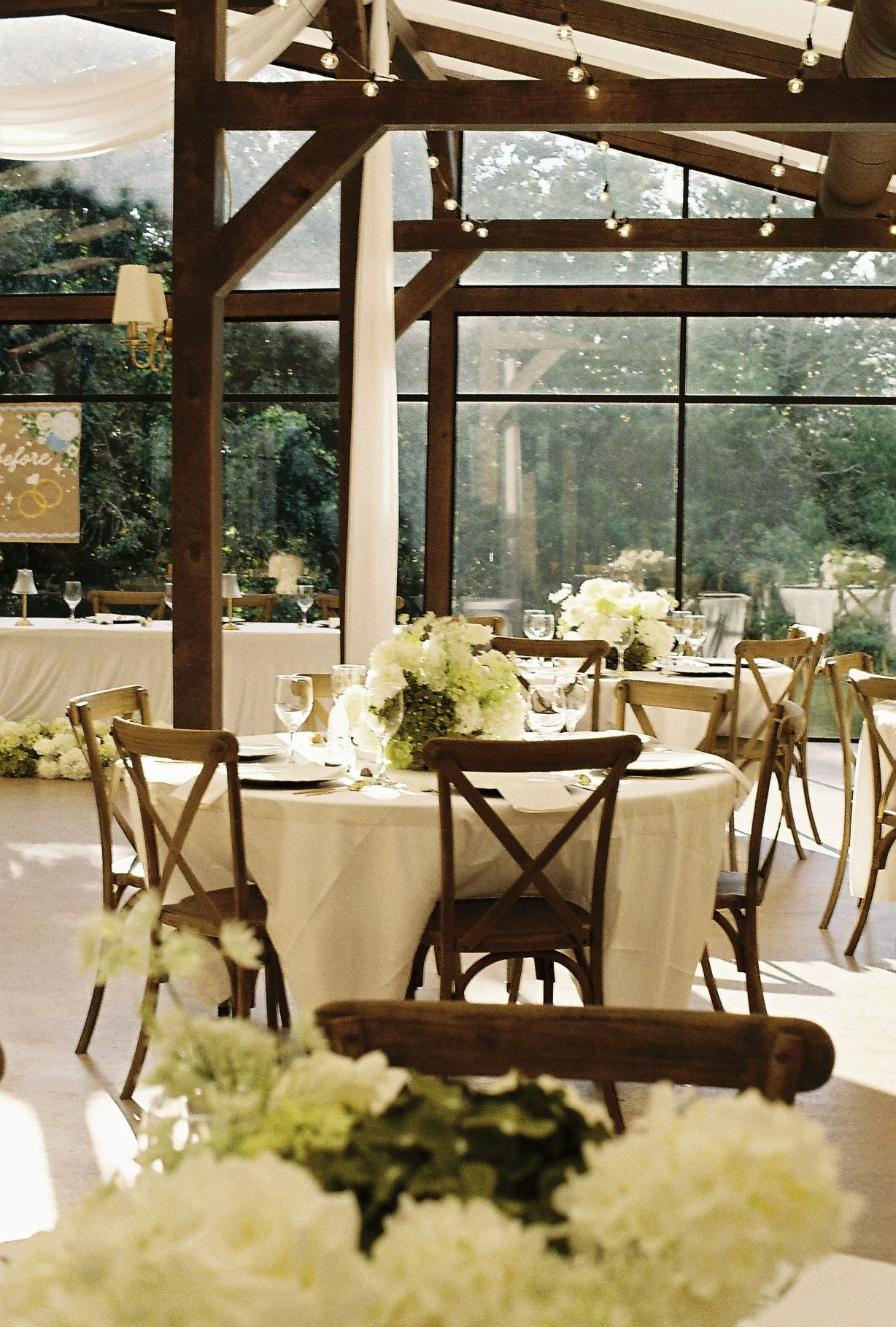 Elegant indoor wedding reception setup with round tables decorated with white tablecloths, floral centerpieces, and glassware, in a venue with large windows and wooden beams decorated with string lights.