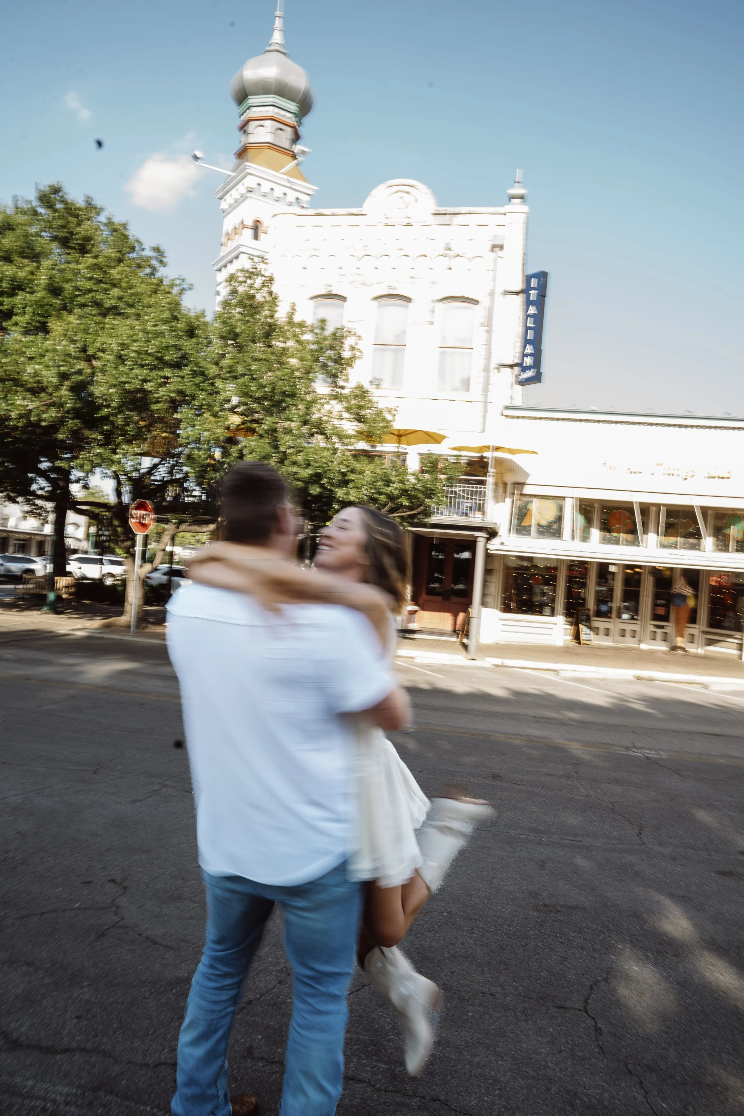 A couple dancing on a city street during the daytime, with the woman wearing a white dress and the man in a white shirt and blue jeans, with a white building and trees in the background.