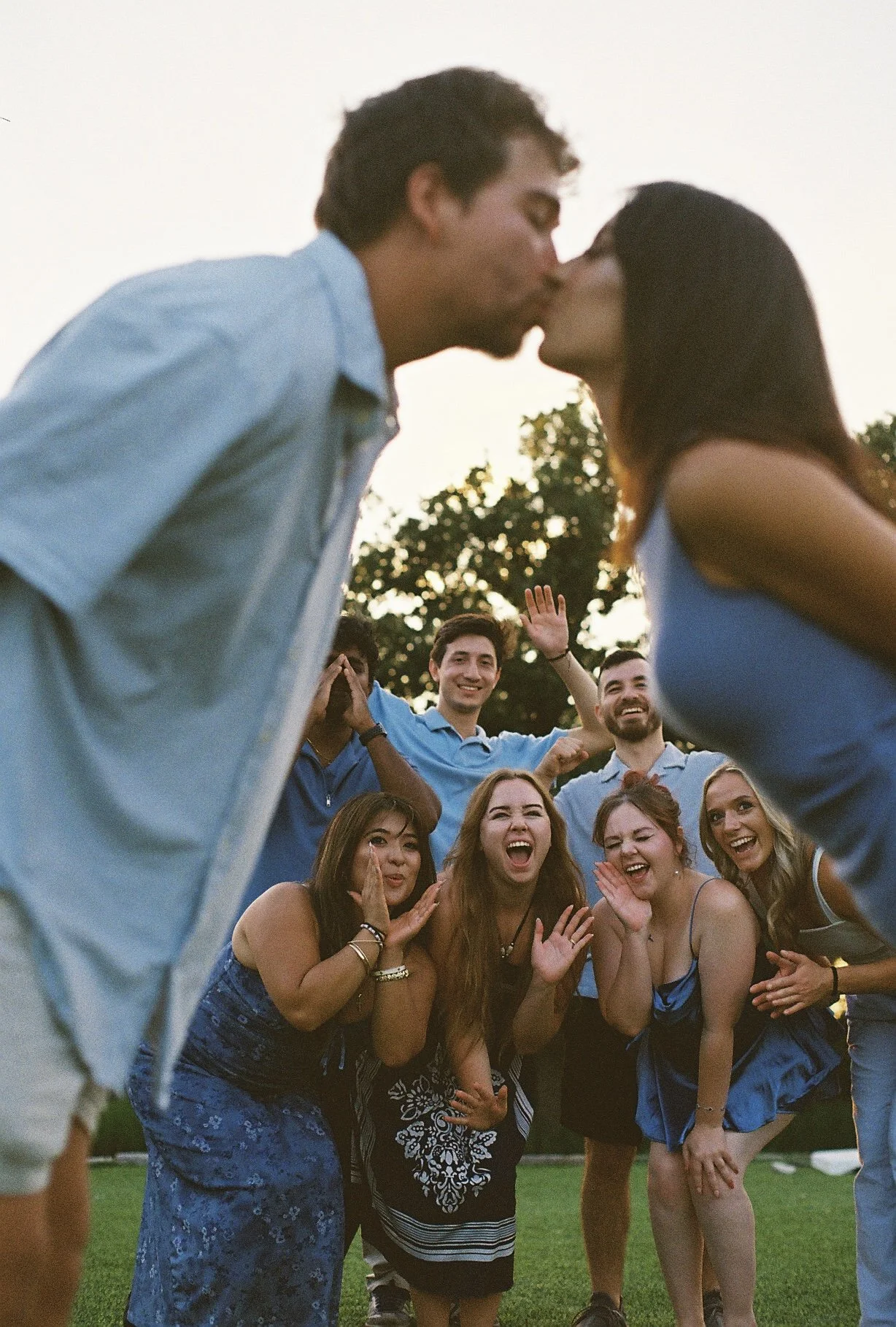 Group of young adults outdoors at sunset, kissing a couple in the foreground, all smiling, with trees in the background.