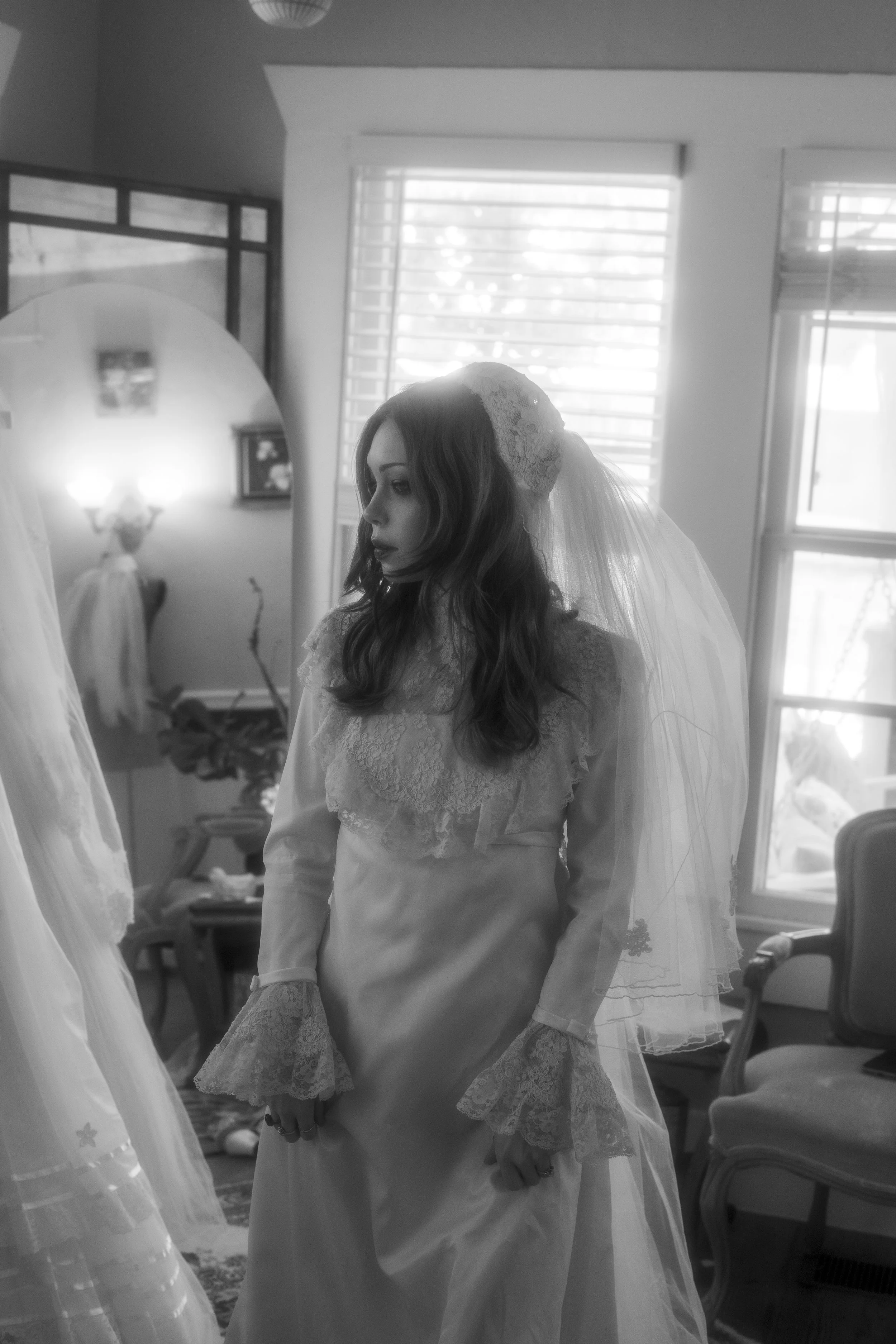 A bride in a lace wedding dress and veil stands indoors near a window, looking down with a contemplative expression.