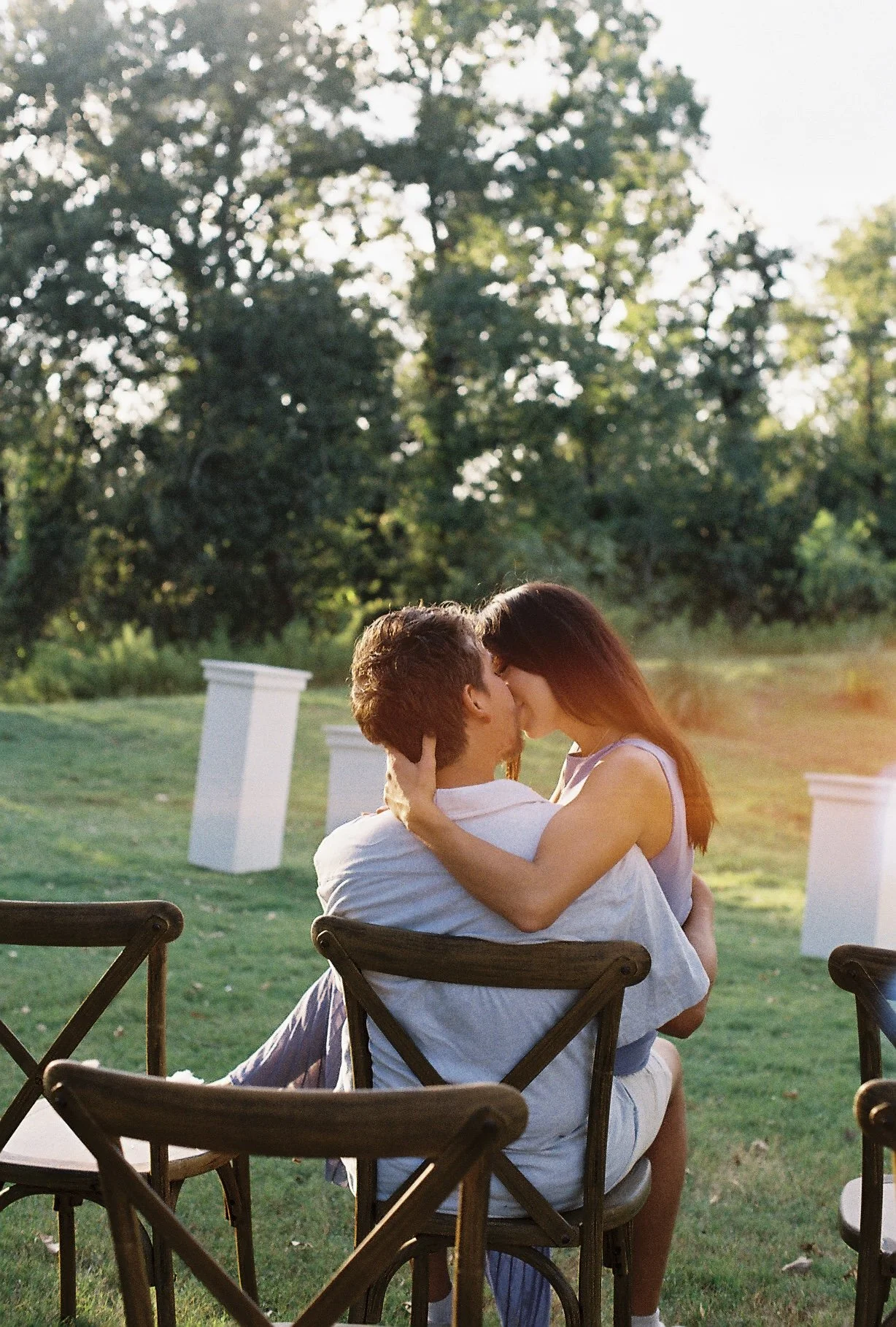 A couple sharing a kiss outdoors during sunset, sitting on chairs at a garden or park with trees in the background.