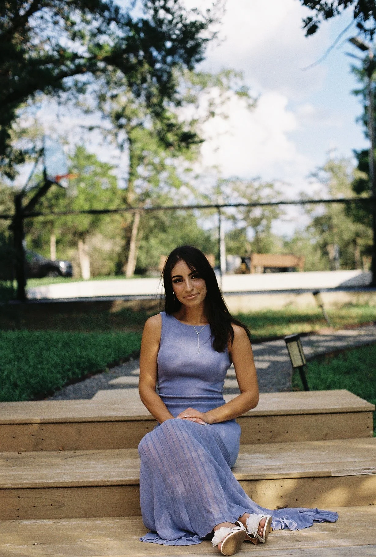 A young woman with dark hair and a sleeveless blue top sitting on wooden steps in a park, with trees and a basketball hoop in the background.