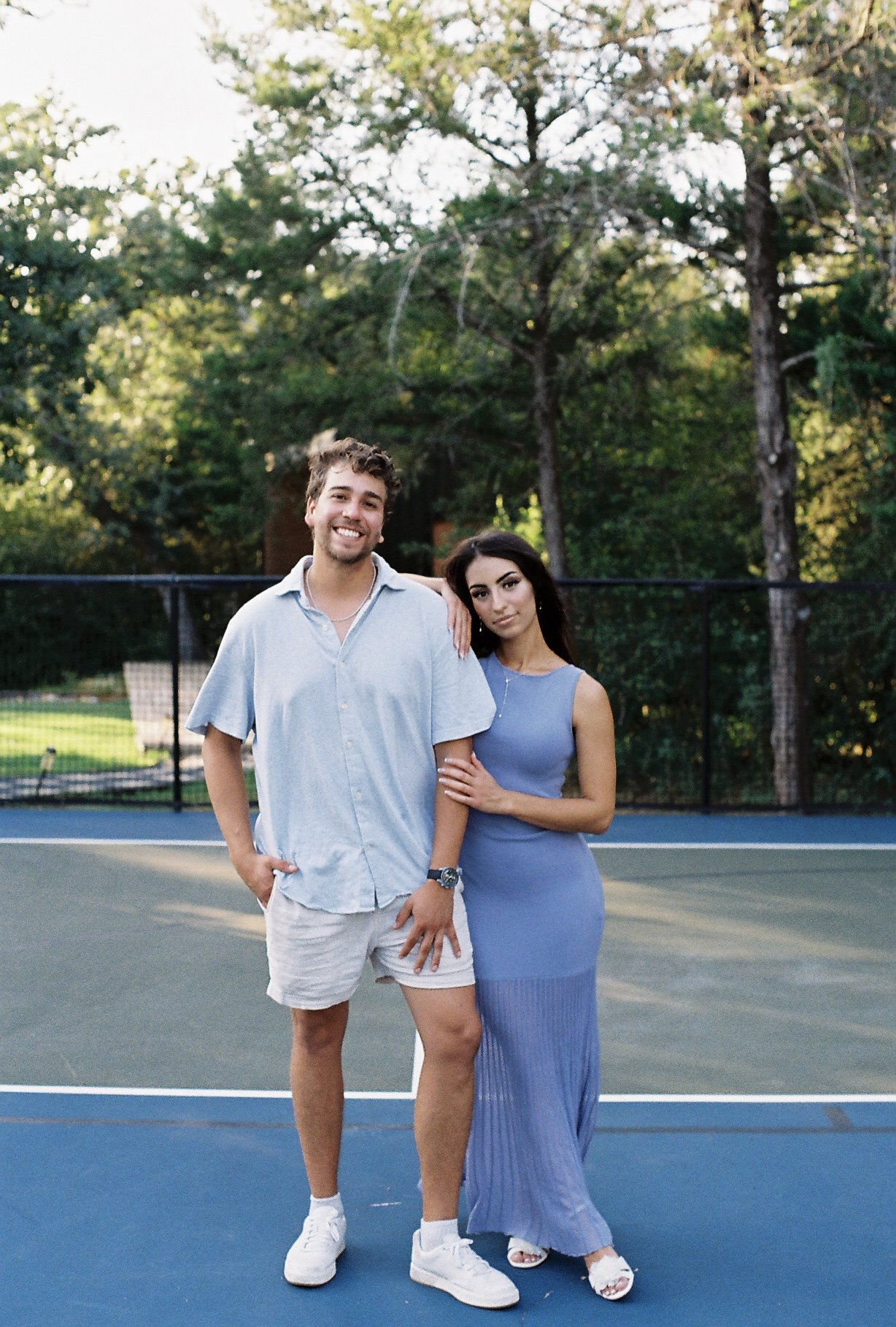 A young man and woman standing on a tennis court, with trees in the background. The man has curly hair, is smiling, and is wearing a light blue short-sleeve shirt, beige shorts, and white sneakers. The woman has long dark hair, a neutral expression, 