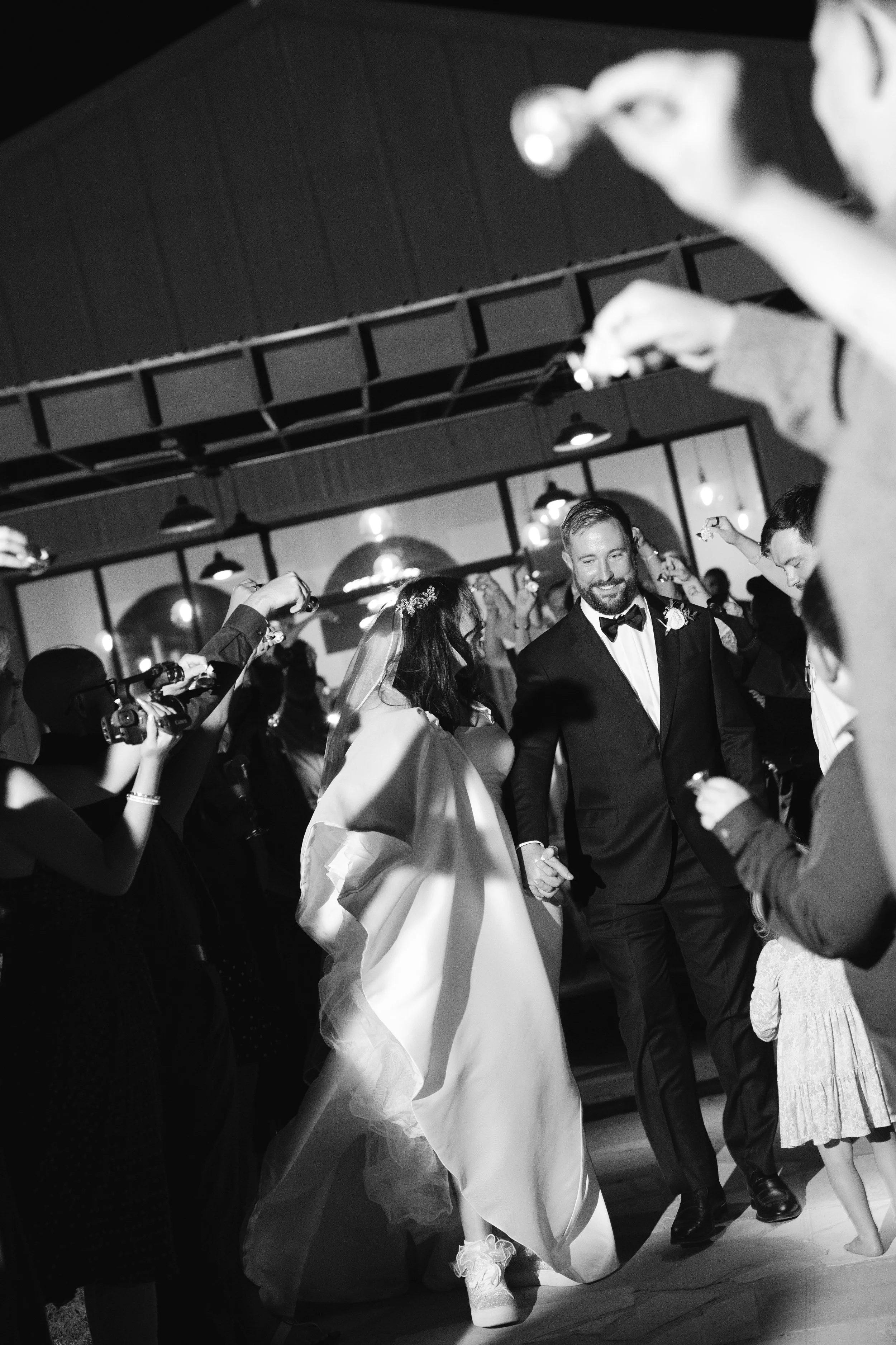 A black and white photo of a wedding reception, with the bride and groom dancing surrounded by guests, some holding cameras and taking pictures, in a room with pendant lighting and a high ceiling.