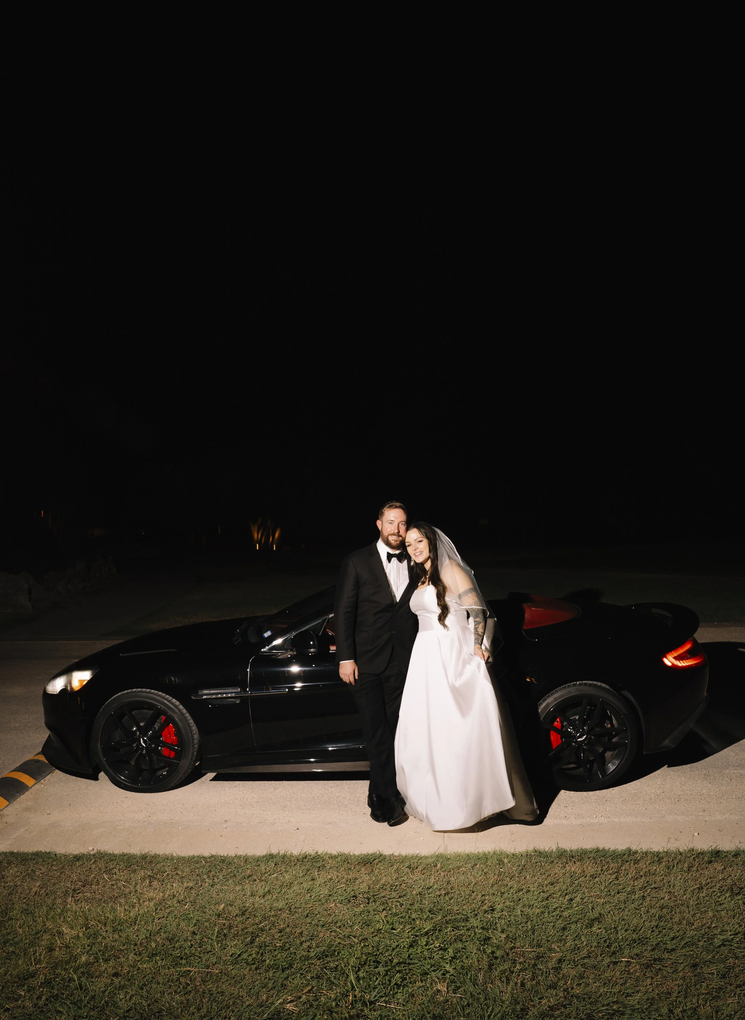 A newlywed couple standing together in front of a black sports car at night, smiling for the camera. The groom is dressed in a black tuxedo and the bride in a white wedding gown with a veil.