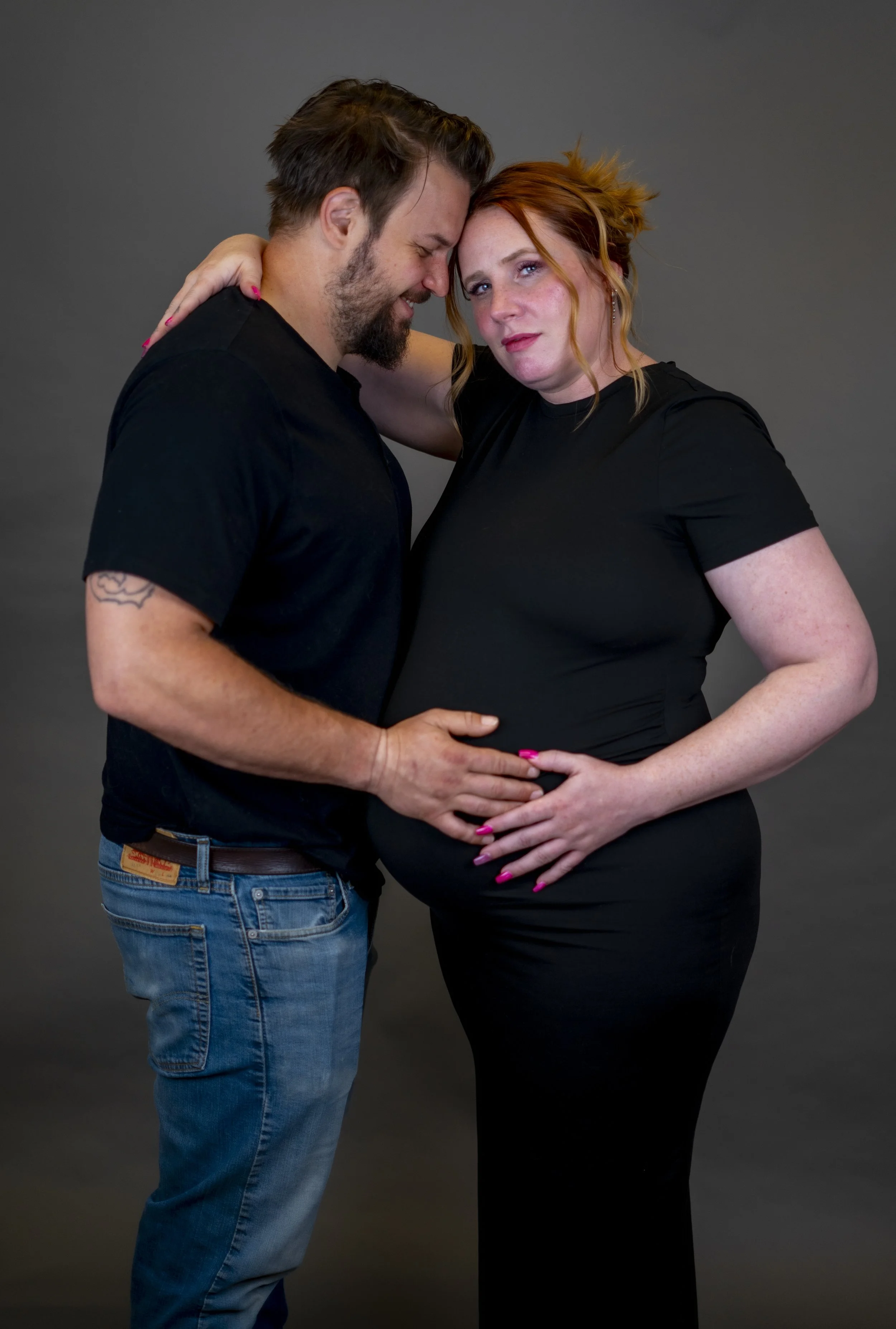 A couple, with the man hugging the pregnant woman, both dressed in black, against a plain gray background.