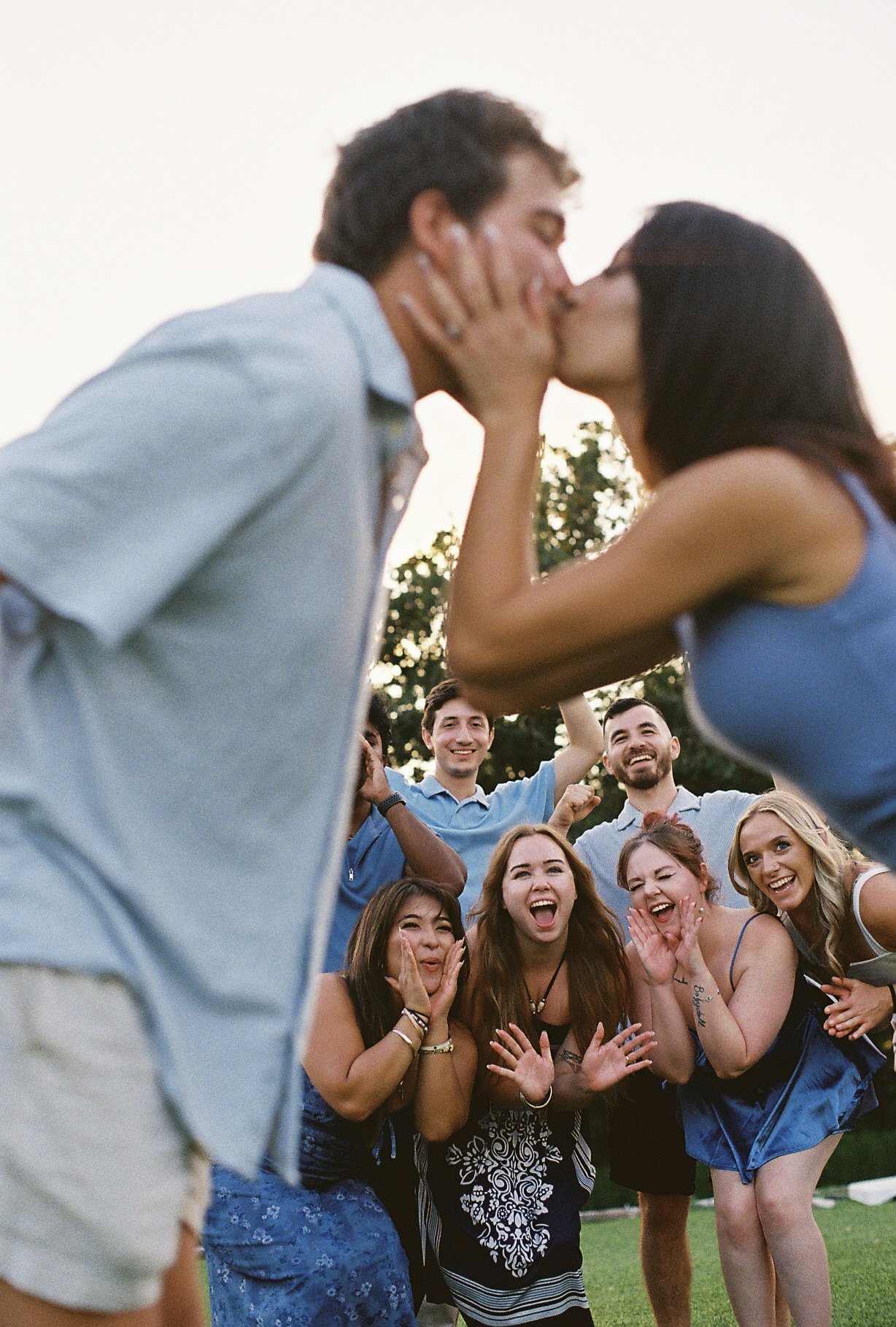 Group of young adults outdoors celebrating, with a couple sharing a kiss in the foreground, others smiling and clapping, during a sunny daytime.
