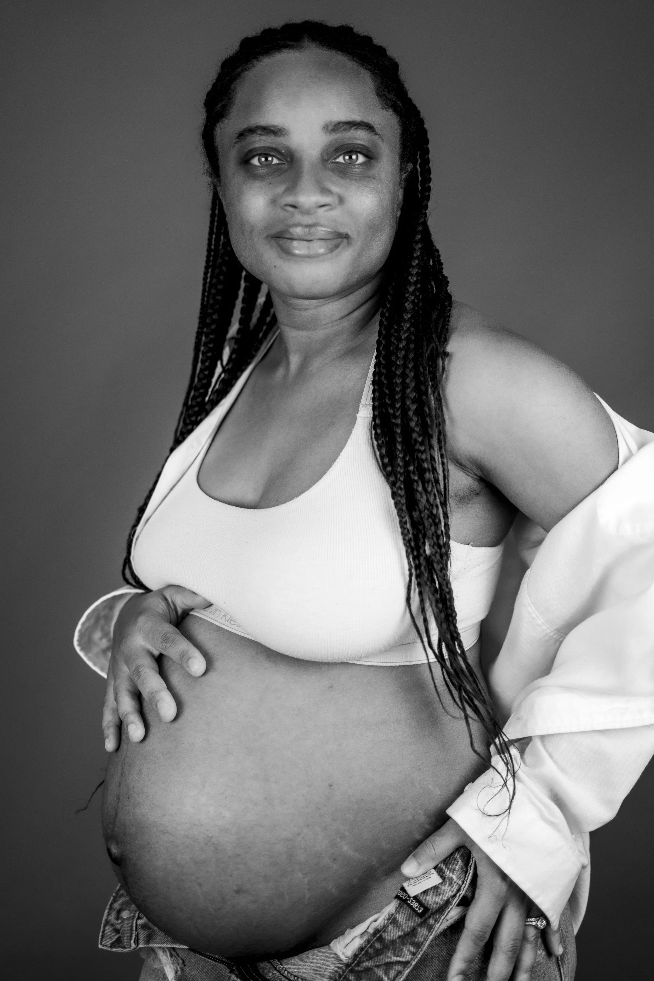 A pregnant woman with long braided hair wears a white sports bra, standing with her hand on her belly and a slight smile, against a plain backdrop.