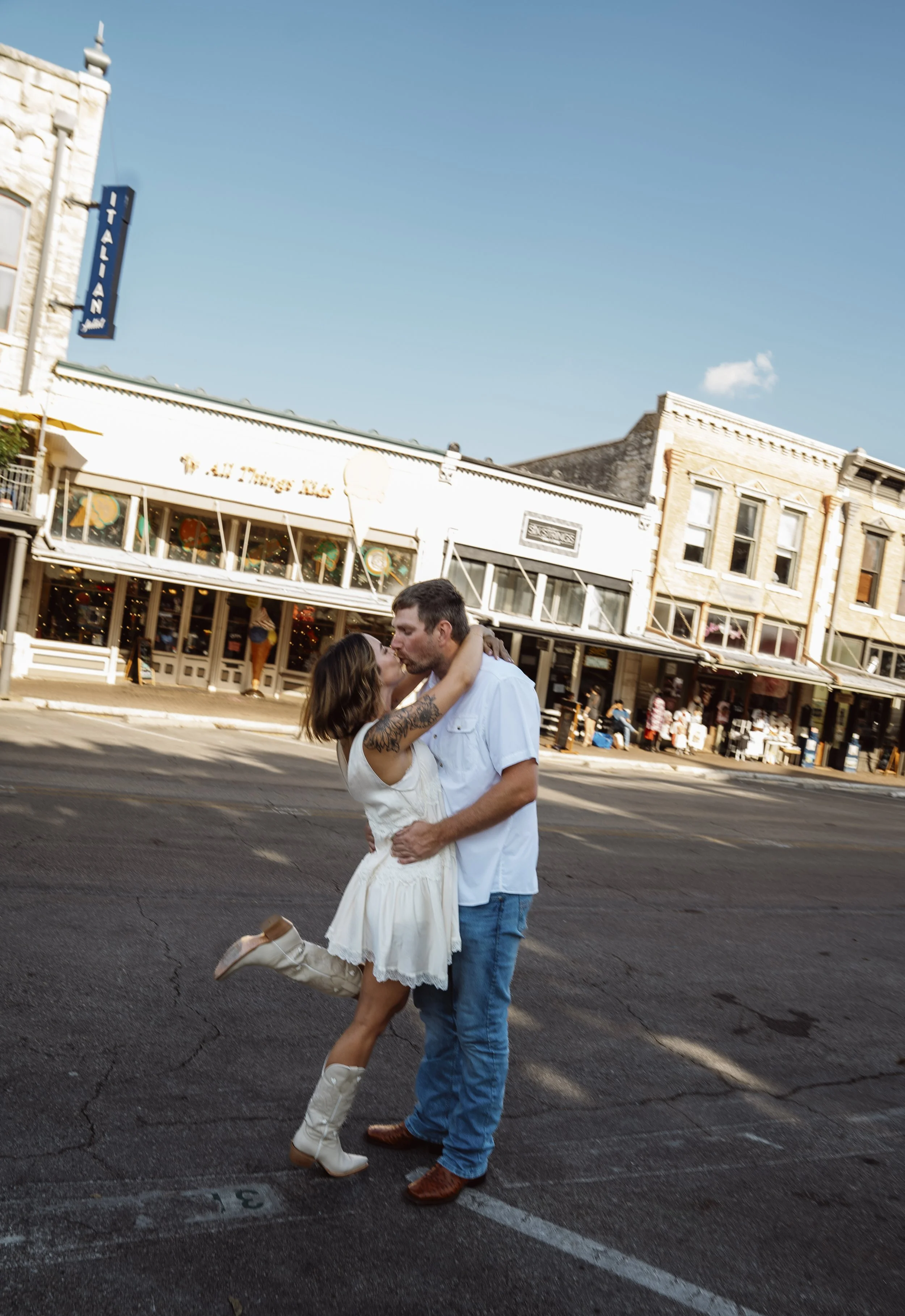 A couple dancing closely in a street in front of shops. The woman is wearing a white dress and cowboy boots, and the man is dressed in a light blue shirt and jeans. The woman has her arms around the man's neck and the man has his hands on her waist. 