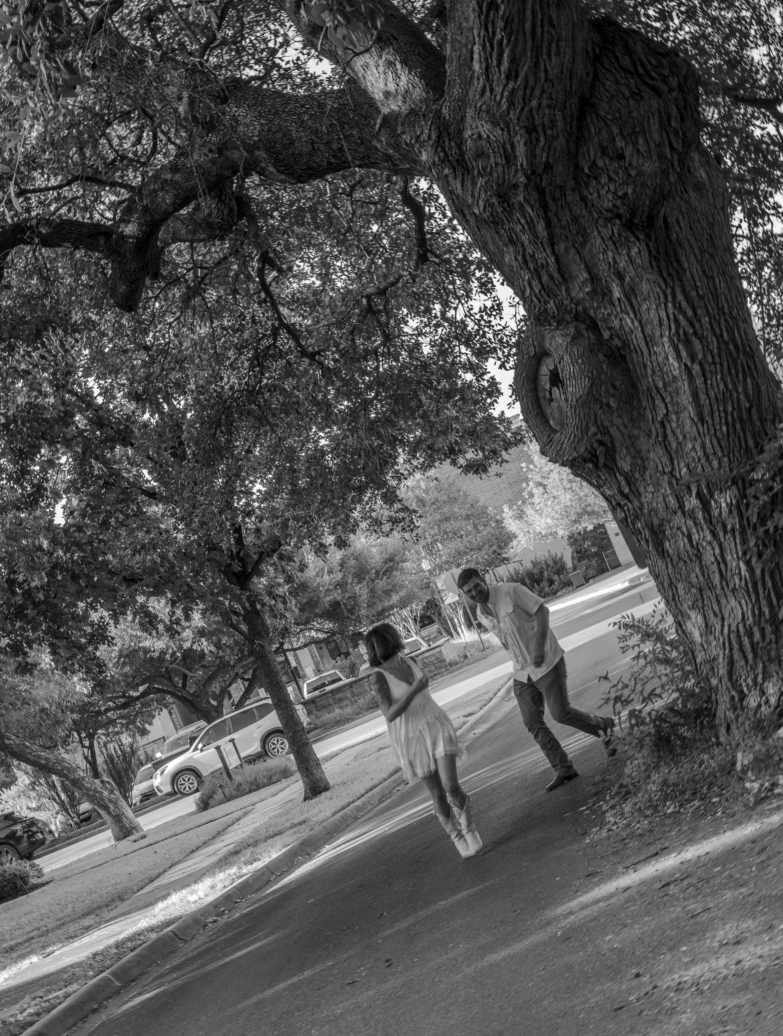 Two people, a man and a woman, walking and talking on a sidewalk beneath a large tree with thick branches and dense leaves. Several cars are parked along the street, and a building is visible in the background.