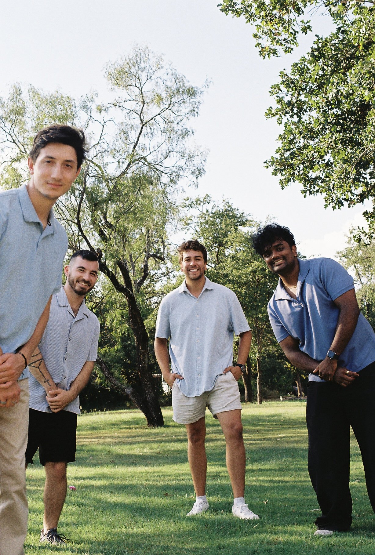 Group of five young men standing outdoors on grass field with trees in background, smiling at the camera, wearing casual summer clothing.