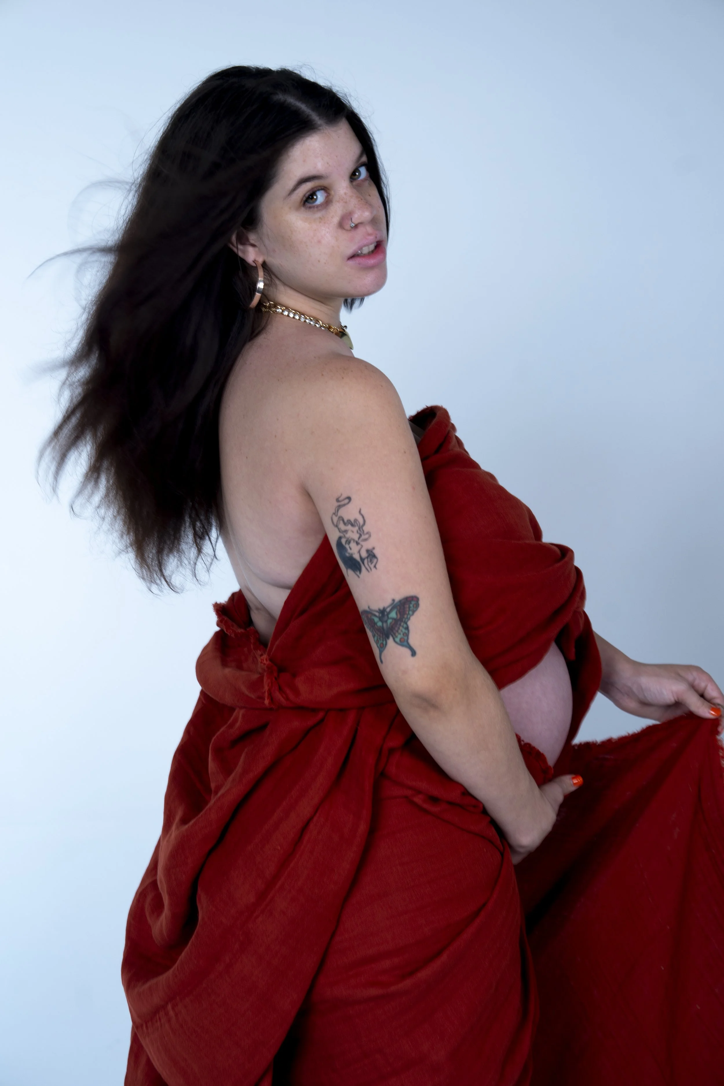 A woman with dark hair, tattoos on her arm, and jewelry, wearing a loose, off-shoulder red dress against a plain background.