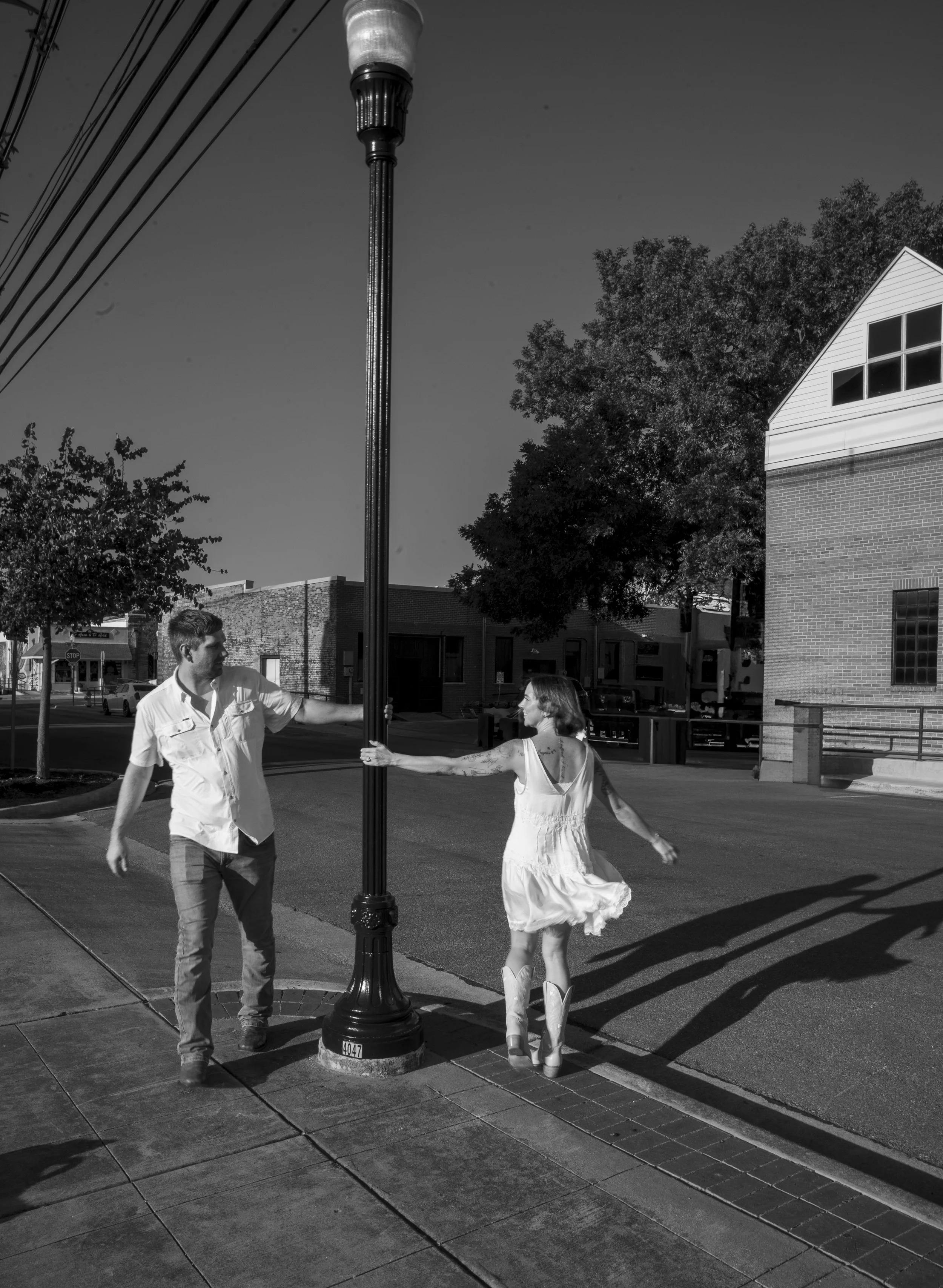 A black and white photo of a man and woman dancing in the street near a lamppost, with a building and trees in the background.
