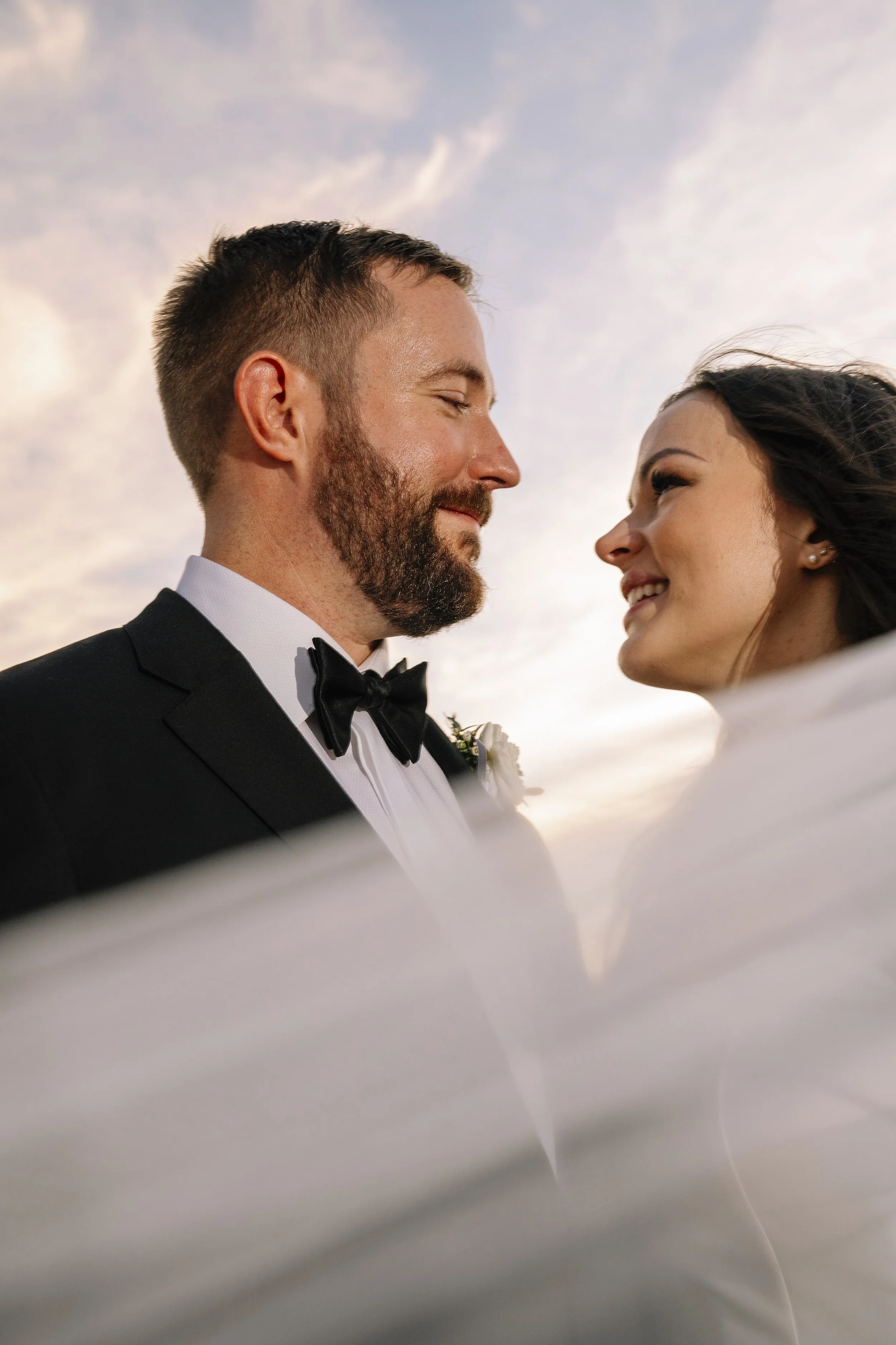 A close-up of a groom and bride facing each other, smiling, during their wedding, with a cloudy sky in the background.
