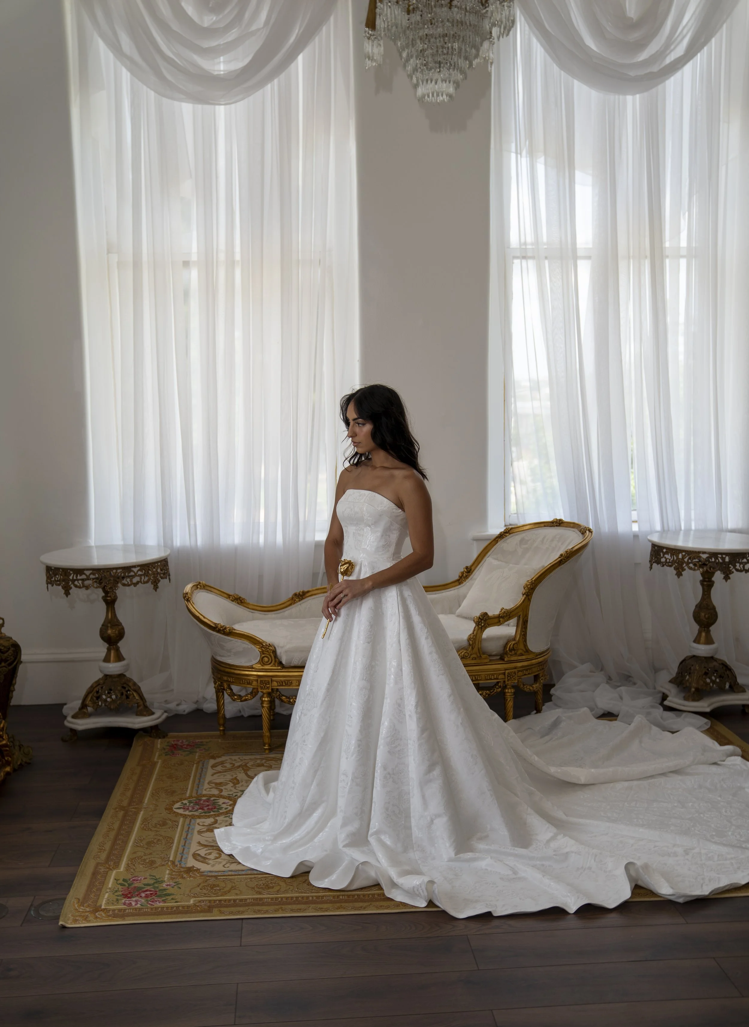 A woman in a white wedding dress standing in a room with tall windows, sheer curtains, and vintage furniture. The dress has a long train spread on the ground.