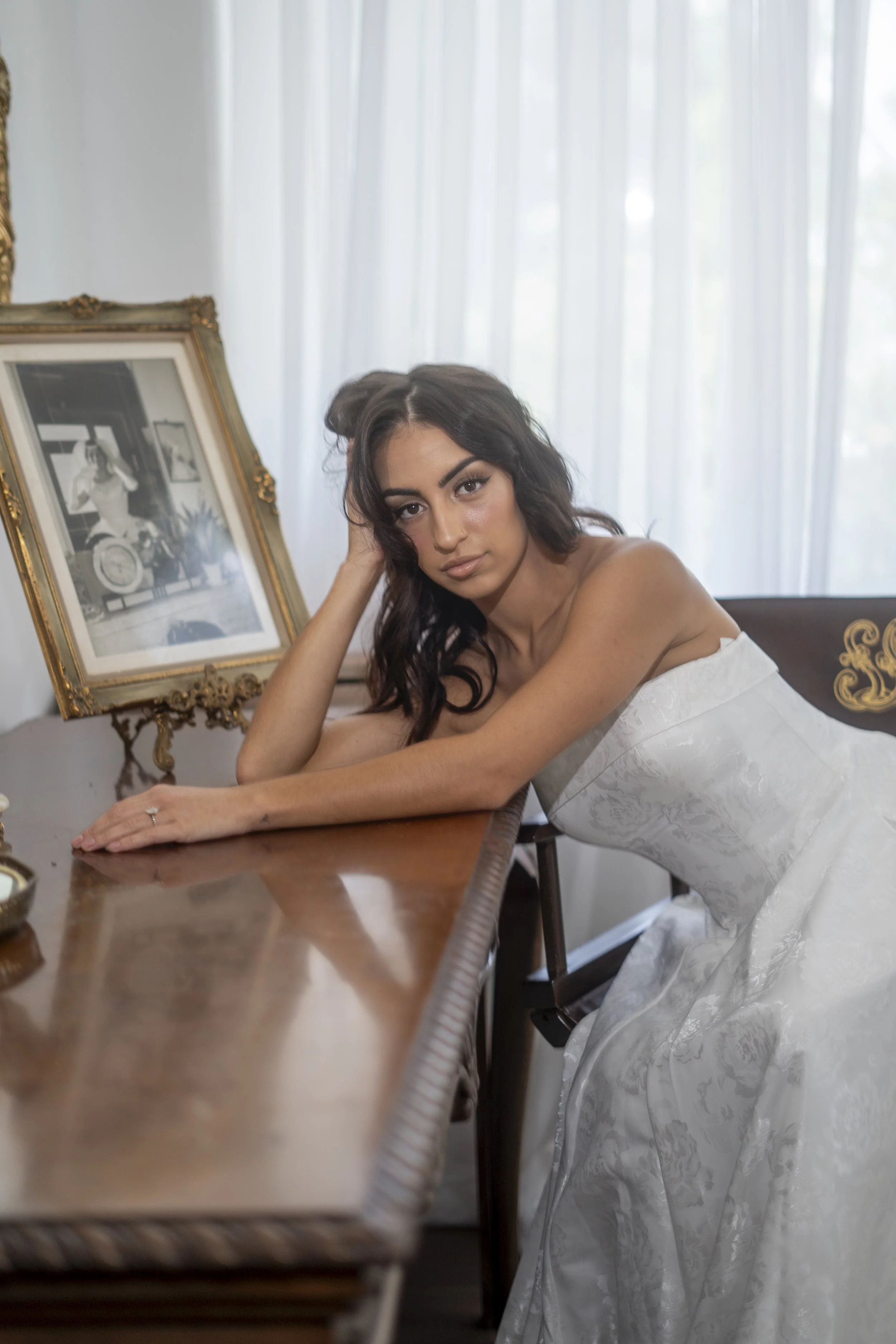 A young woman in a white wedding dress leaning on a wooden table, with her left arm resting on the table and her hand supporting her head. She has dark hair and is looking at the camera. Behind her, there are white curtains and a framed black-and-whi