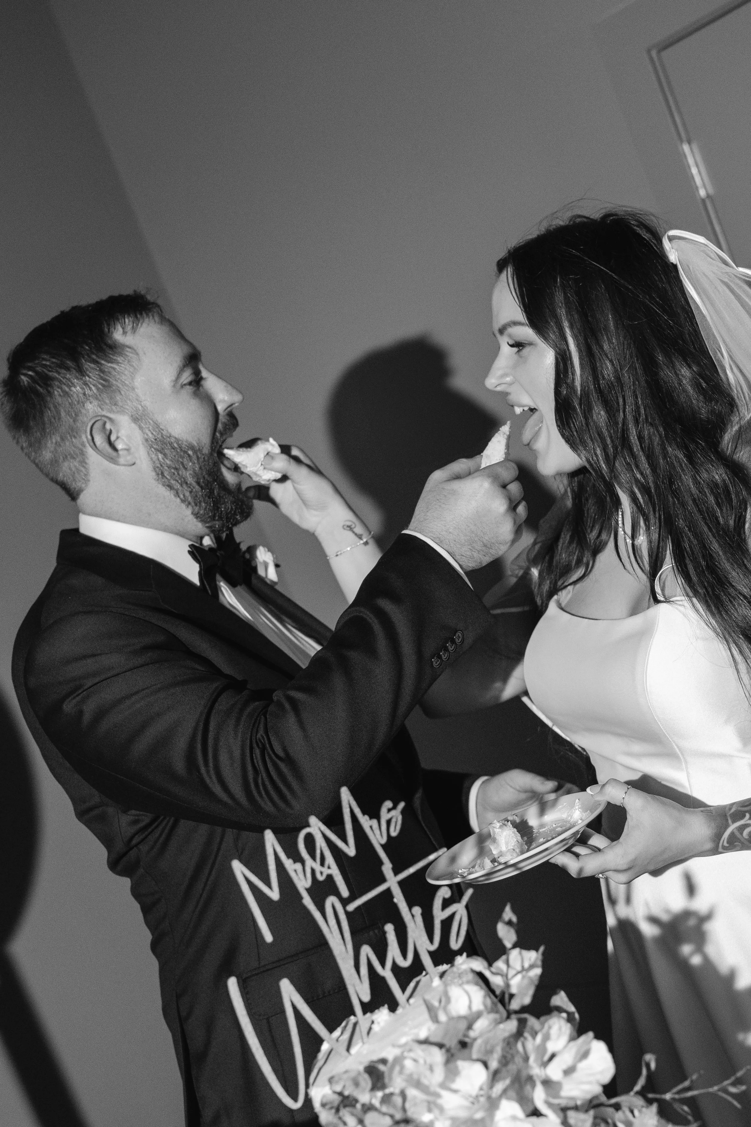 A man in a tuxedo feeds a woman in a wedding dress cake at a wedding reception. The woman is holding a plate of cake and smiling.