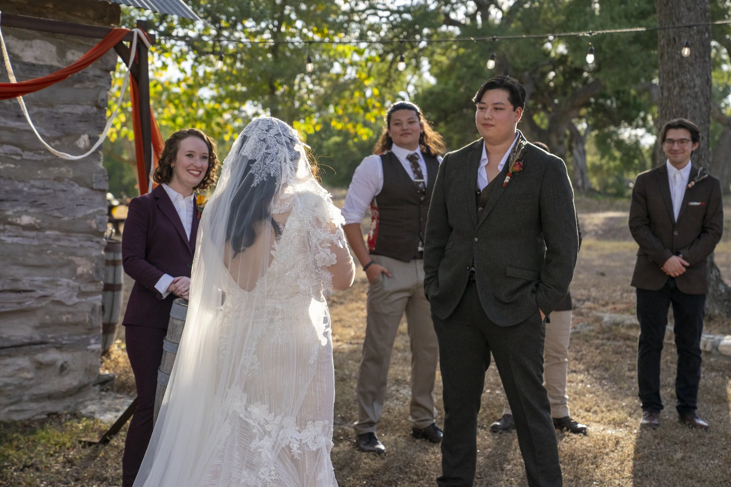 A wedding ceremony outdoors with a bride in a white dress and lace veil, and a groom in a dark suit, looking at each other. Three other people, including two women and one man, stand in the background watching. The scene is set among trees with sunlight filtering through.