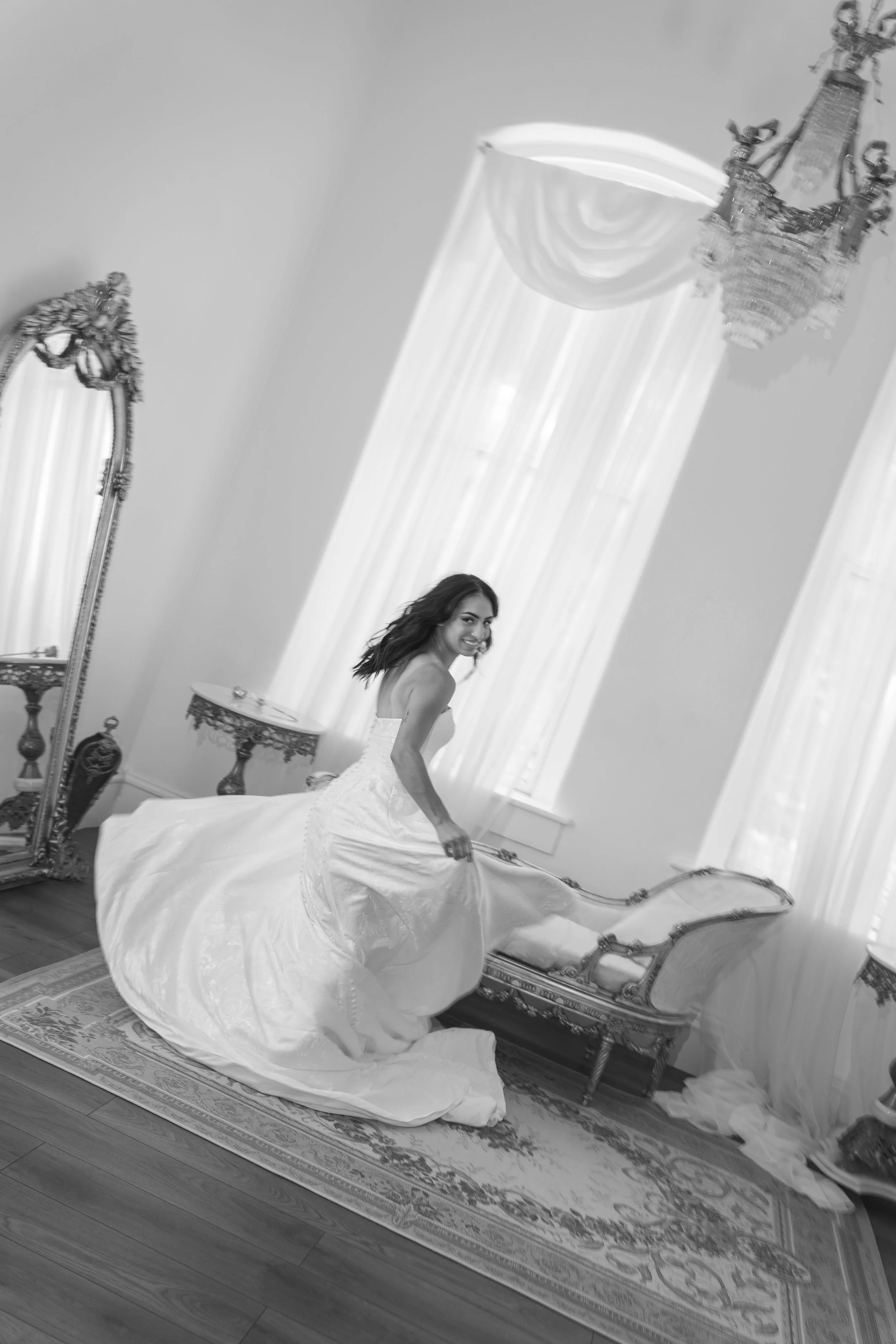 A woman in a wedding gown is kneeling on a patterned rug. The gown is spread out on the floor around her, and she is smiling at the camera. The room has tall windows with curtains, an ornate mirror, and vintage-style furniture.