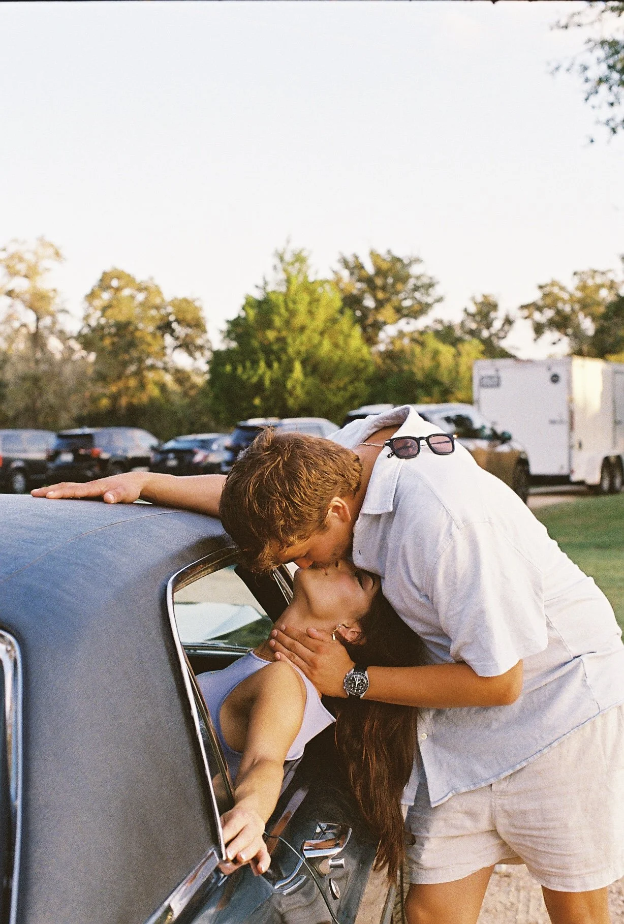 A couple sharing a kiss at the window of a vintage car in a parking lot with trees in the background.