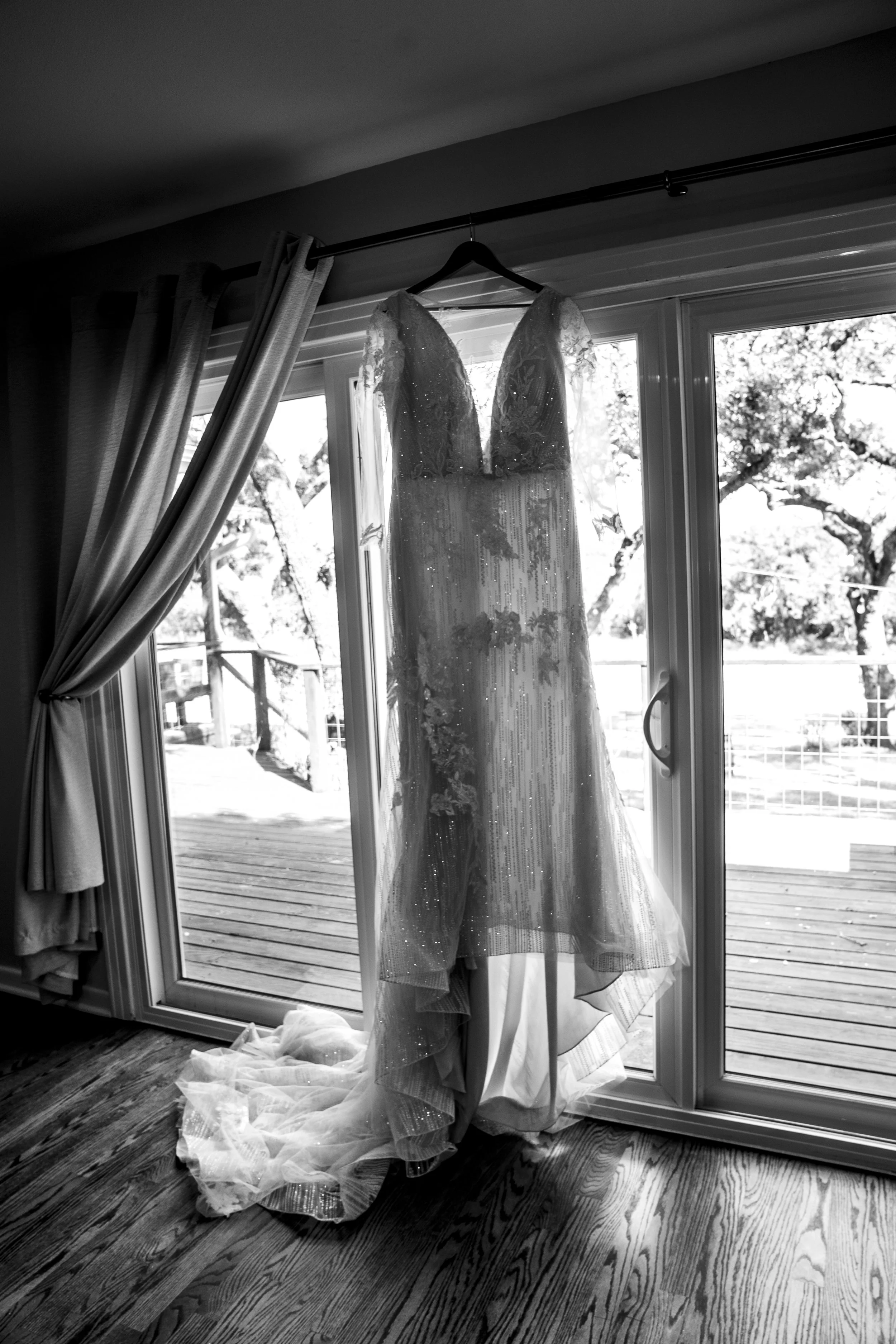 Wedding dress hanging on a hanger in front of sliding glass door, with curtains partially open to reveal outdoor trees and a deck.