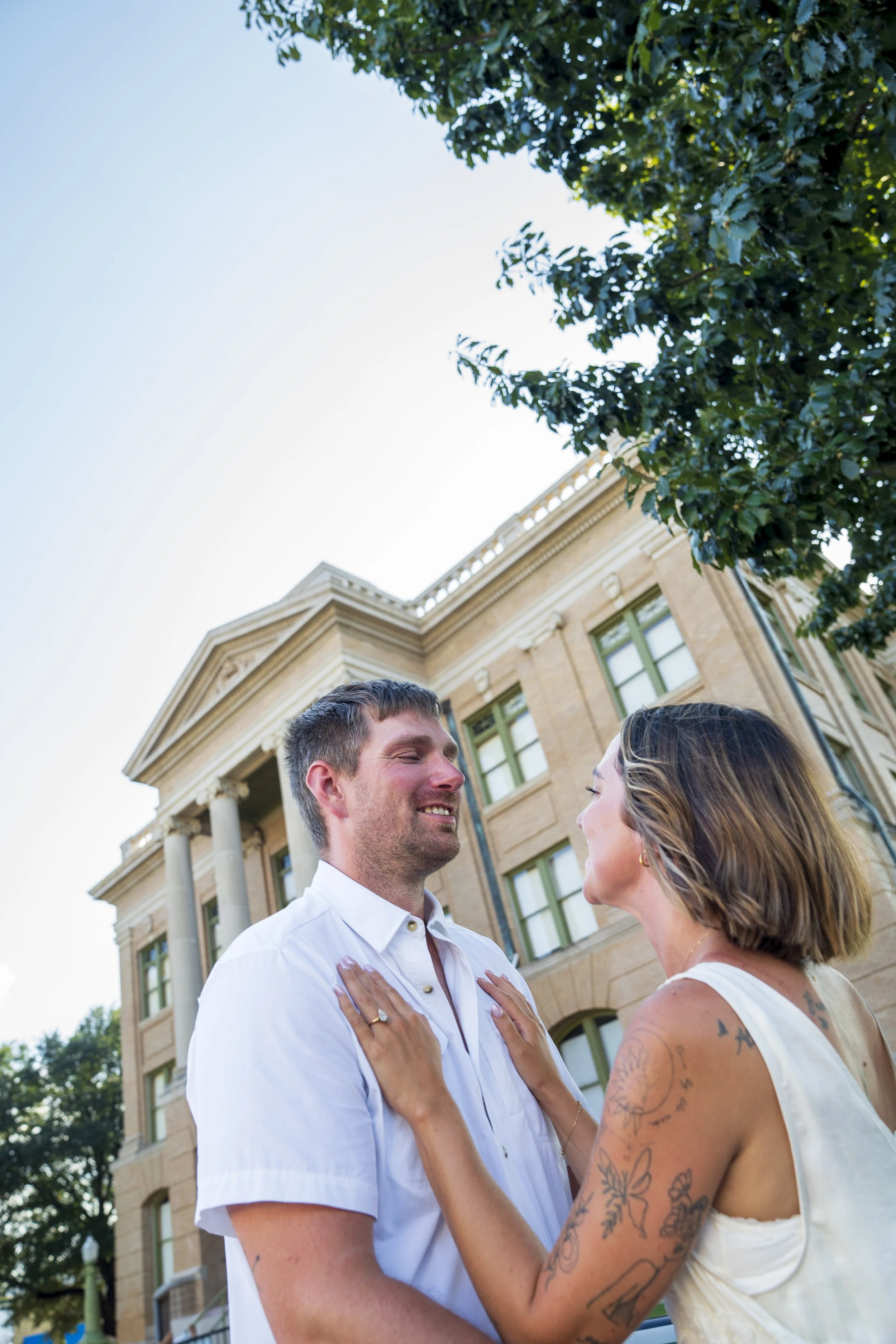 A couple with tattoos gazing at each other outdoors in front of a historic building with columns, green window frames, and trees with green leaves.