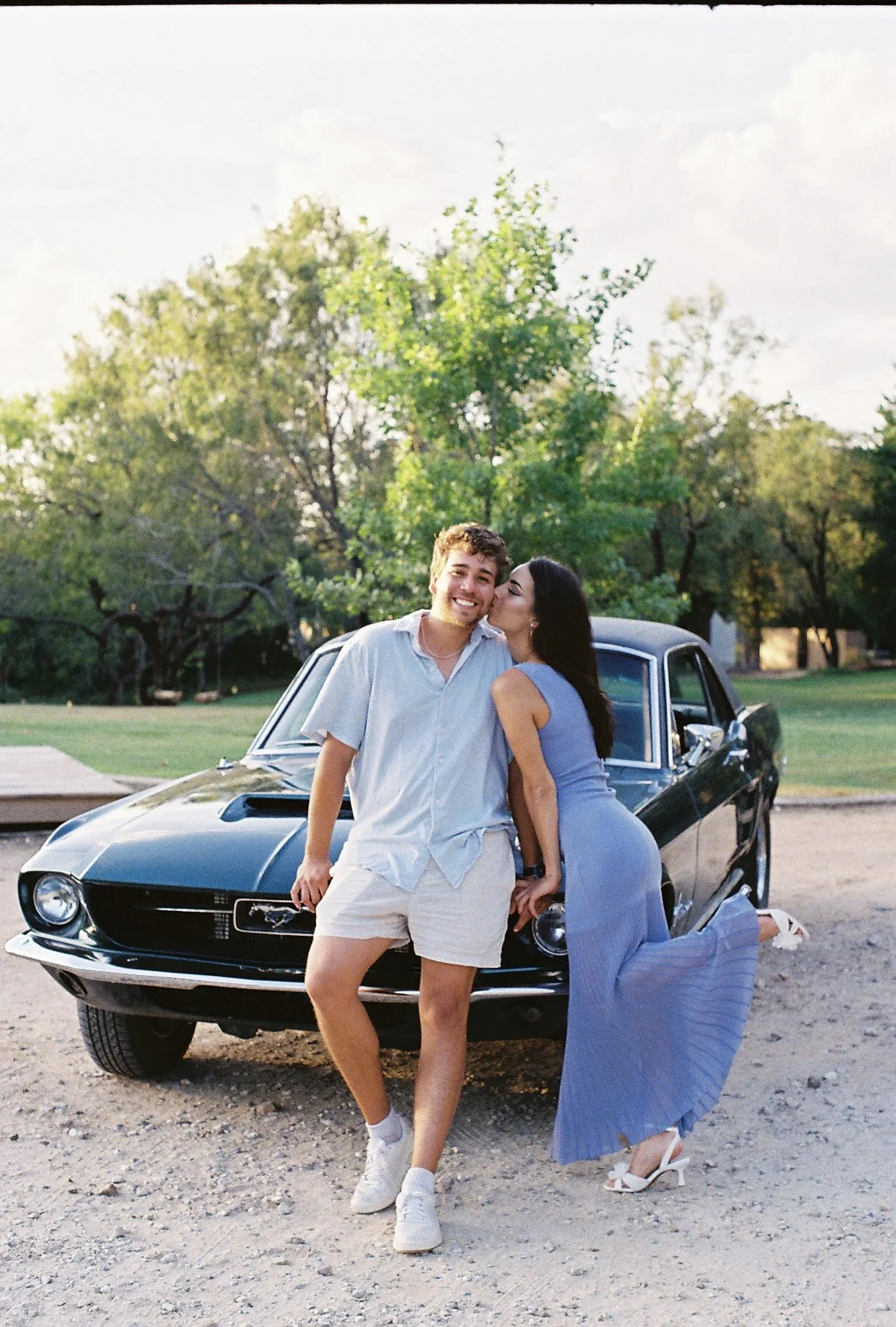 A young couple is posing in front of a black classic Ford Mustang car outdoors, with the woman kissing the man's cheek. They are dressed in summer clothing, and the background features green trees and a partly cloudy sky.