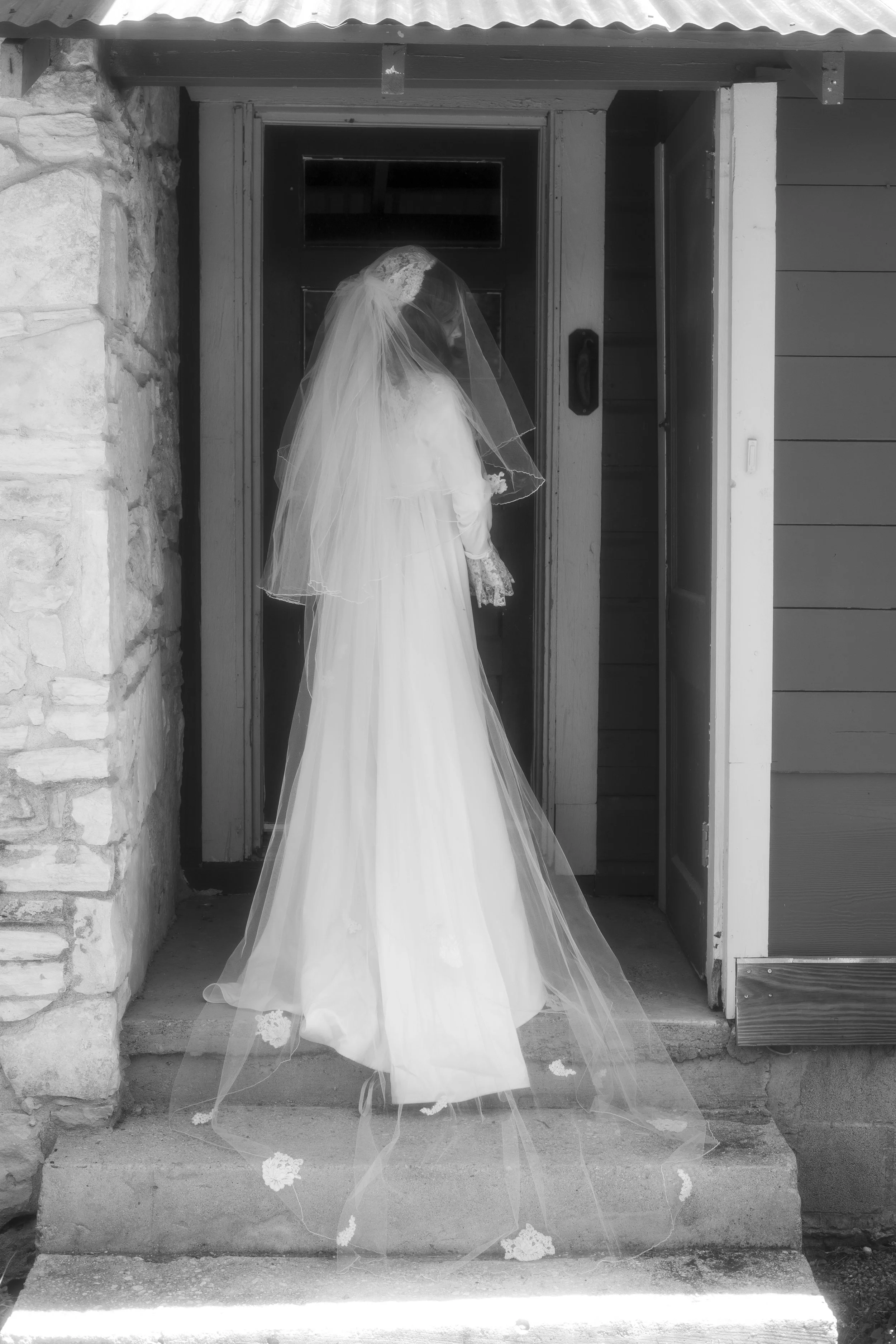 A bride wearing a white wedding dress and veil standing at the entrance of a building, with stone and wood siding around the doorway, viewed in black and white.