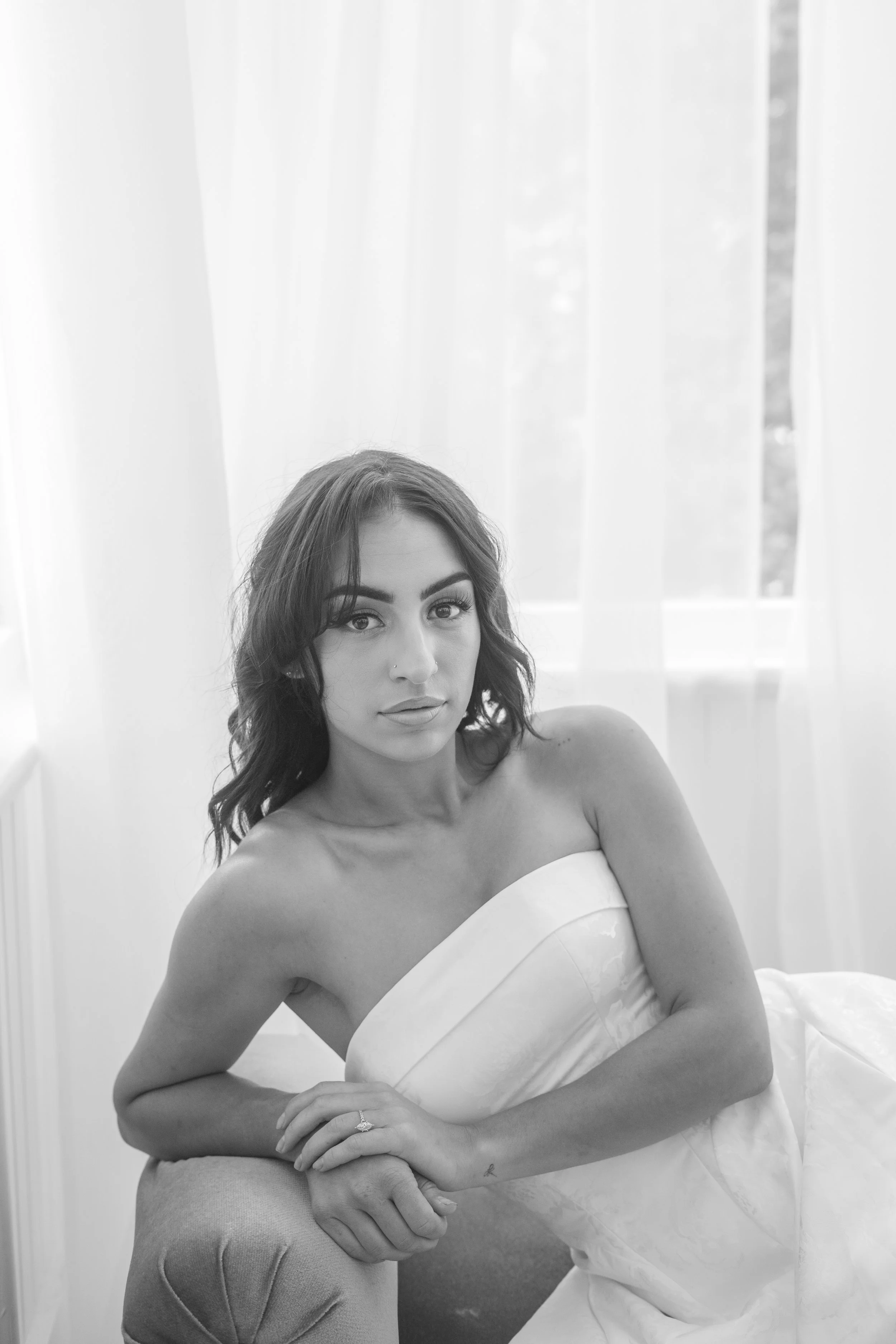 Black and white photo of a woman with dark hair, wearing a strapless dress, sitting on a chair with one arm resting on her knee and the other on her lap, in front of a window with light curtains.